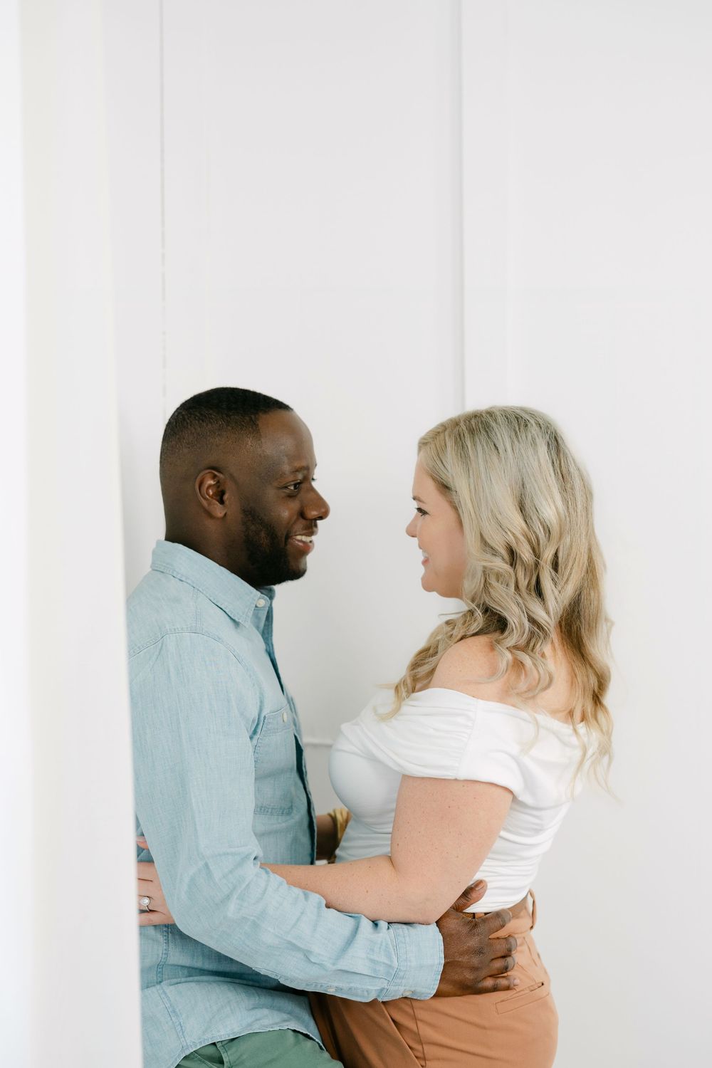 A smiling couple embraces in a bright white room, the man in a light blue button-up shirt and the woman in a white top and tan pants.