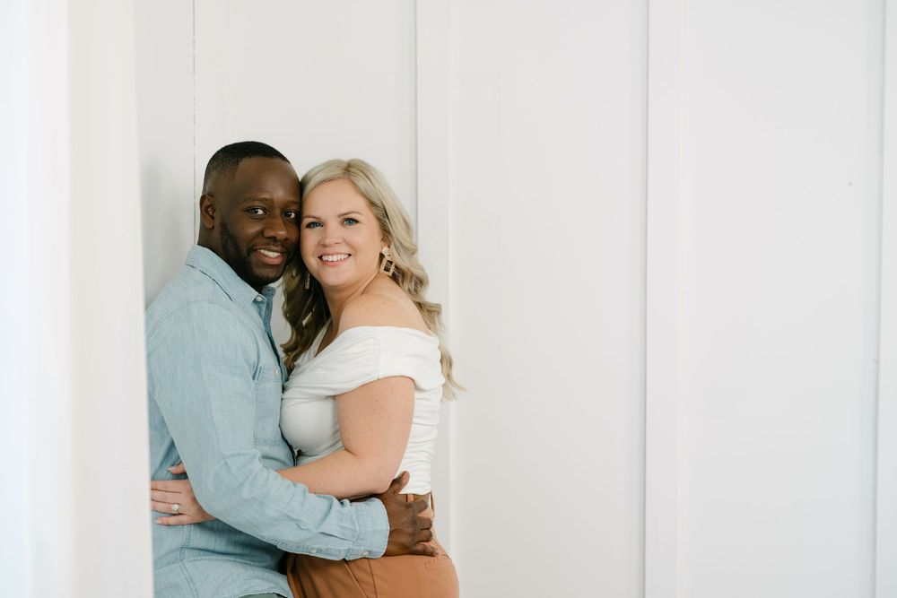 Smiling couple embracing in a bright white room, the man in a light blue button-up shirt and the woman in a white off-shoulder top.