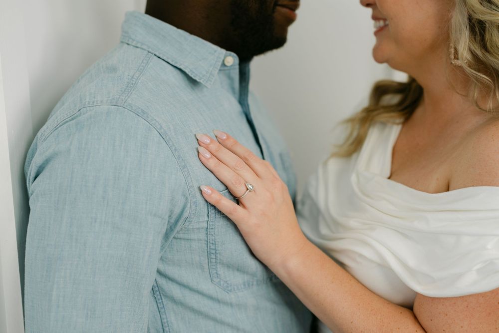Woman with wavy blonde hair and a diamond engagement ring rests her hand on a man's chest in a light blue chambray shirt.