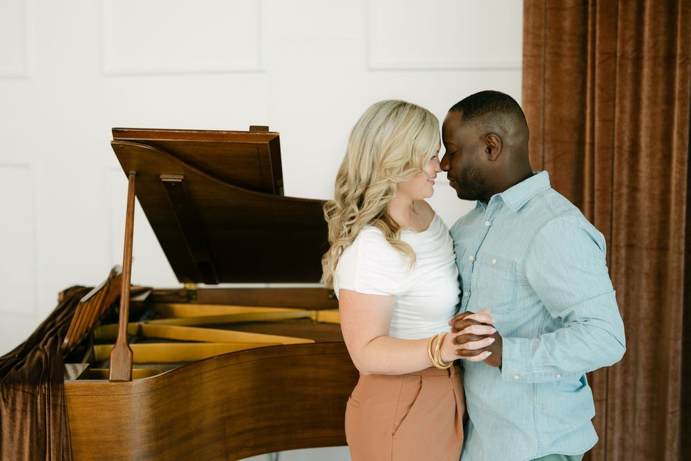 Smiling couple in an embrace beside a grand piano, the woman in a white top and the man in a blue button-up shirt, by brown curtains.