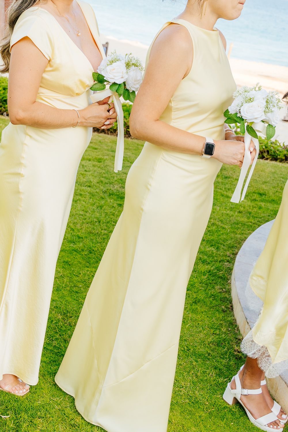 Bridesmaids in pale yellow satin gowns hold white floral bouquets while standing on a lawn beside the ocean.