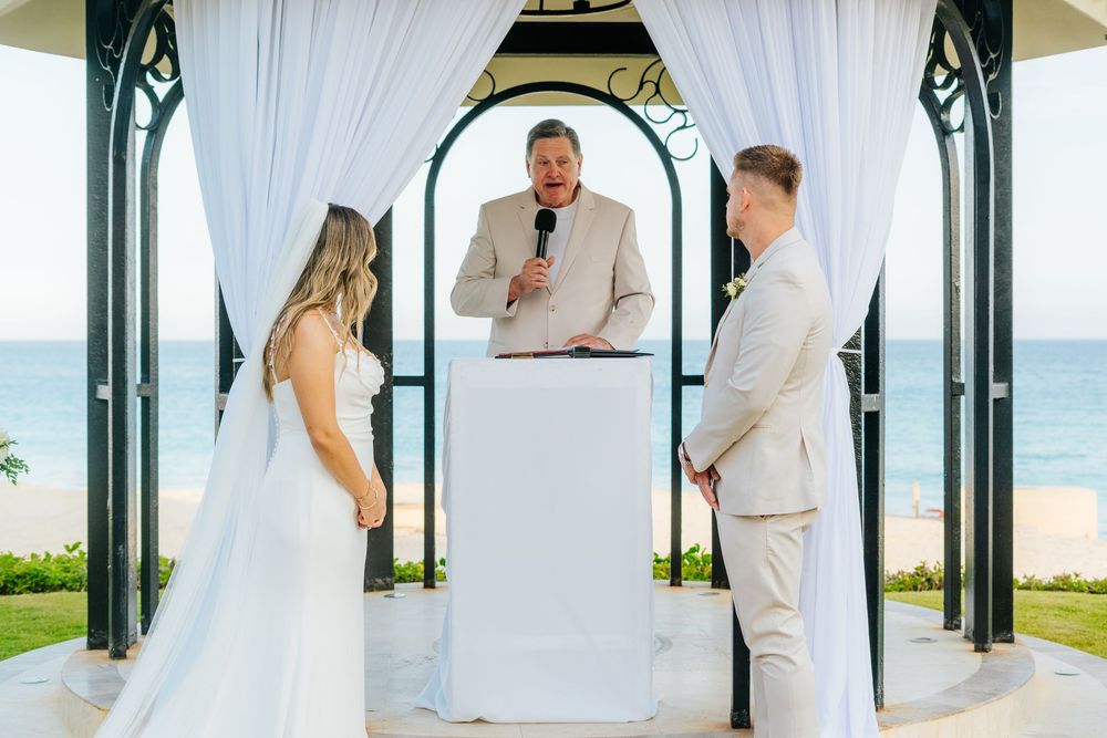 Bride in a white gown and groom in a cream suit exchange vows before an officiant under a white draped gazebo on a beachfront.