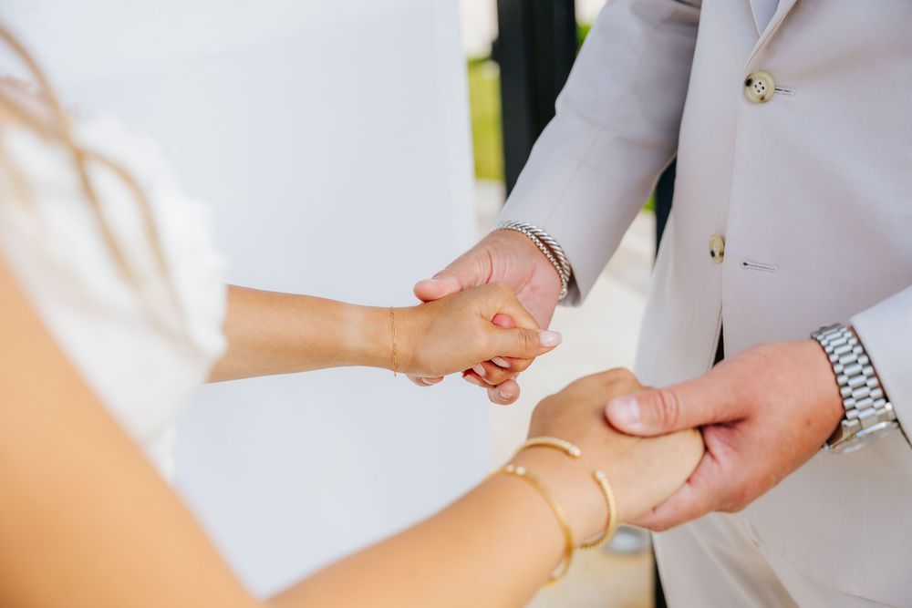 Bride in a white dress and groom in a light gray suit hold hands during a wedding ceremony, her gold bracelets visible.