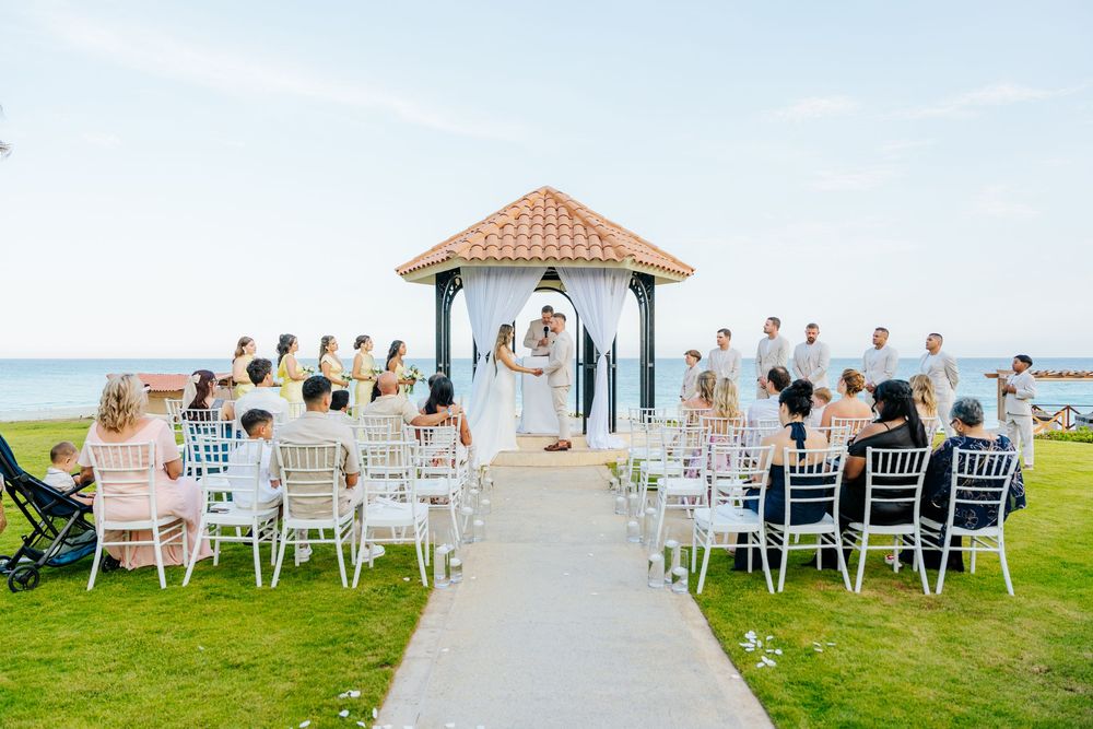 Outdoor beachside wedding ceremony with guests seated on white chairs facing a couple under a gazebo, with the ocean in the background.