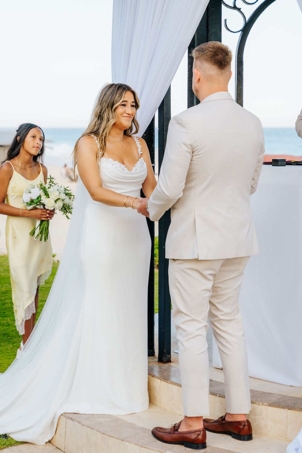 A smiling bride in a white satin gown holds hands with the groom in a beige suit during an outdoor beachside wedding ceremony.