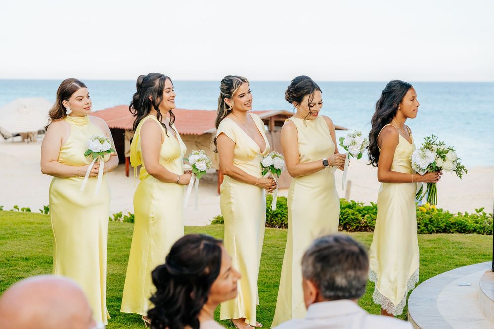 Bridesmaids in flowing pale yellow gowns hold white bouquets during a beachfront wedding ceremony as seated guests look on.