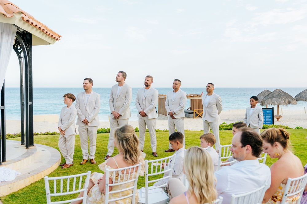 Groomsmen in light linen suits stand in a row on the lawn as guests watch a beachfront wedding ceremony by the ocean.