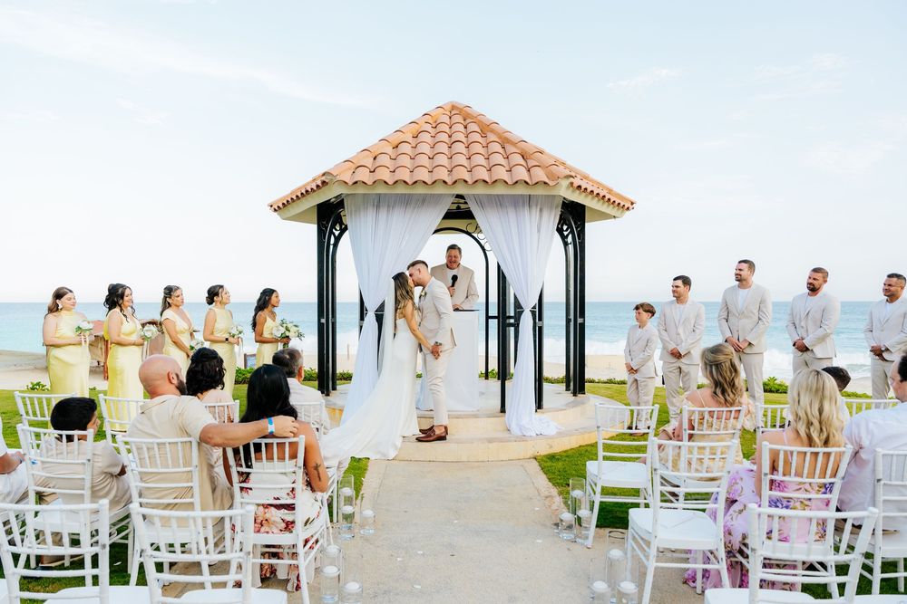 Bride and groom kiss under a white-draped gazebo during a beachside wedding ceremony as guests in white chairs look on.