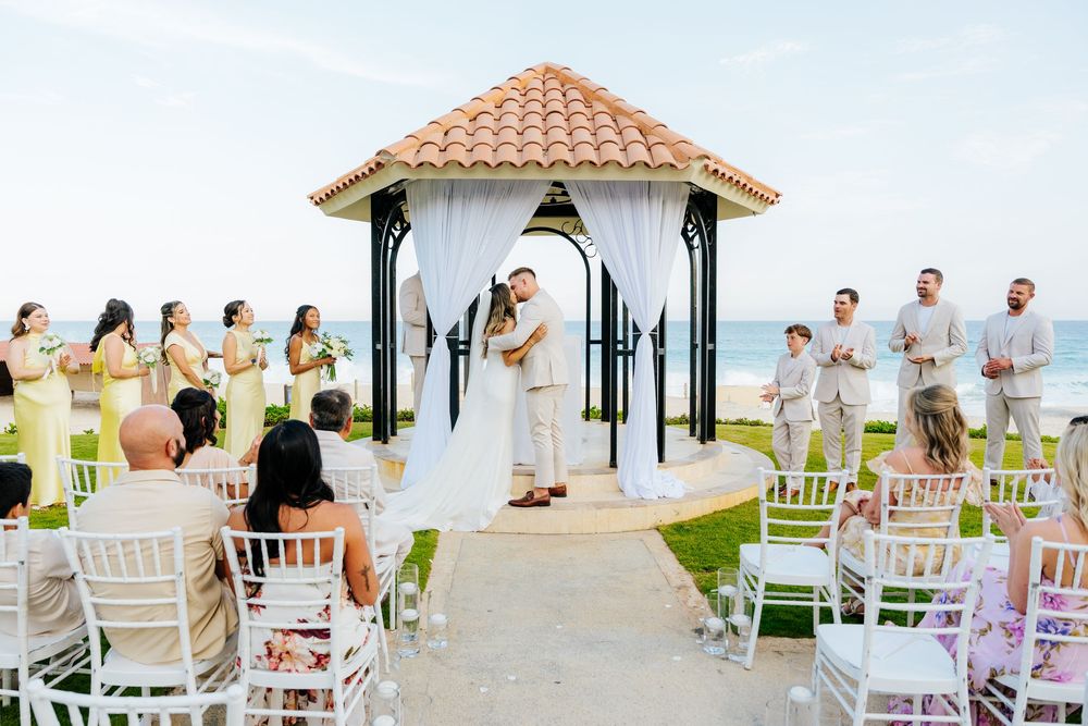 Bride and groom exchange vows under a tiled-roof beachfront gazebo as guests and wedding party look on during an oceanside ceremony.