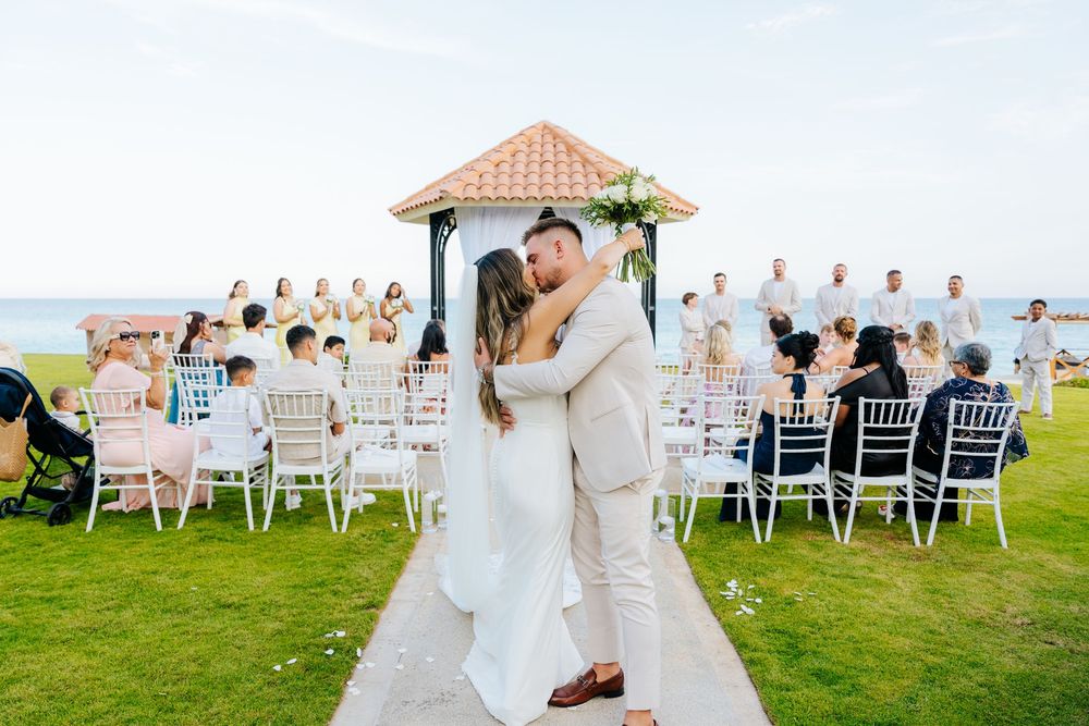 Bride and groom embrace on an aisle between white chairs at an oceanfront wedding ceremony, with guests seated and a gazebo behind them.
