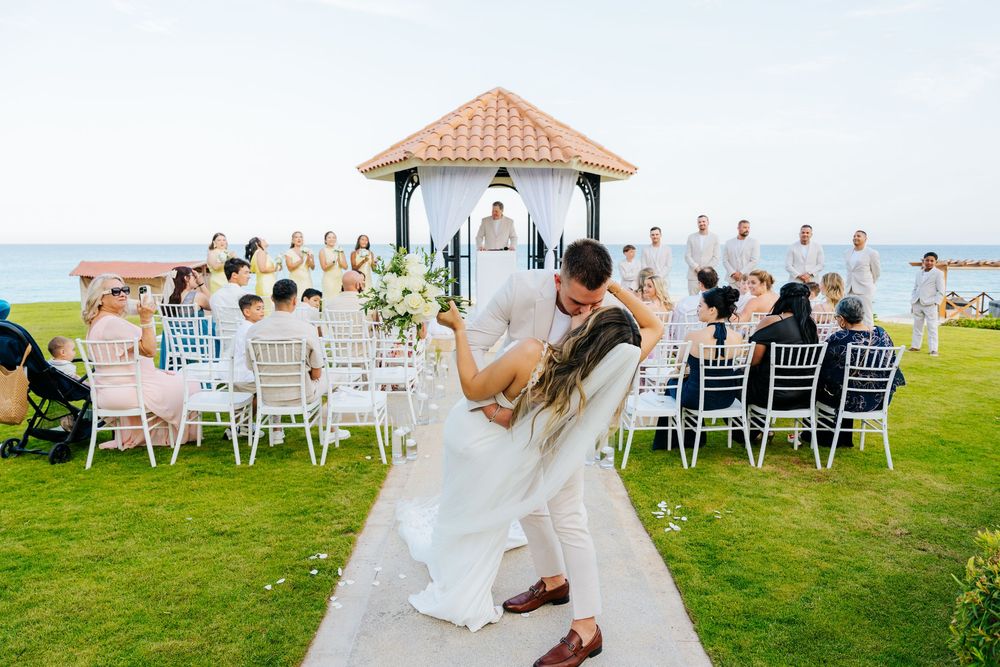 Newlywed couple shares a romantic dip kiss on an aisle at an oceanfront gazebo wedding ceremony as seated guests look on.