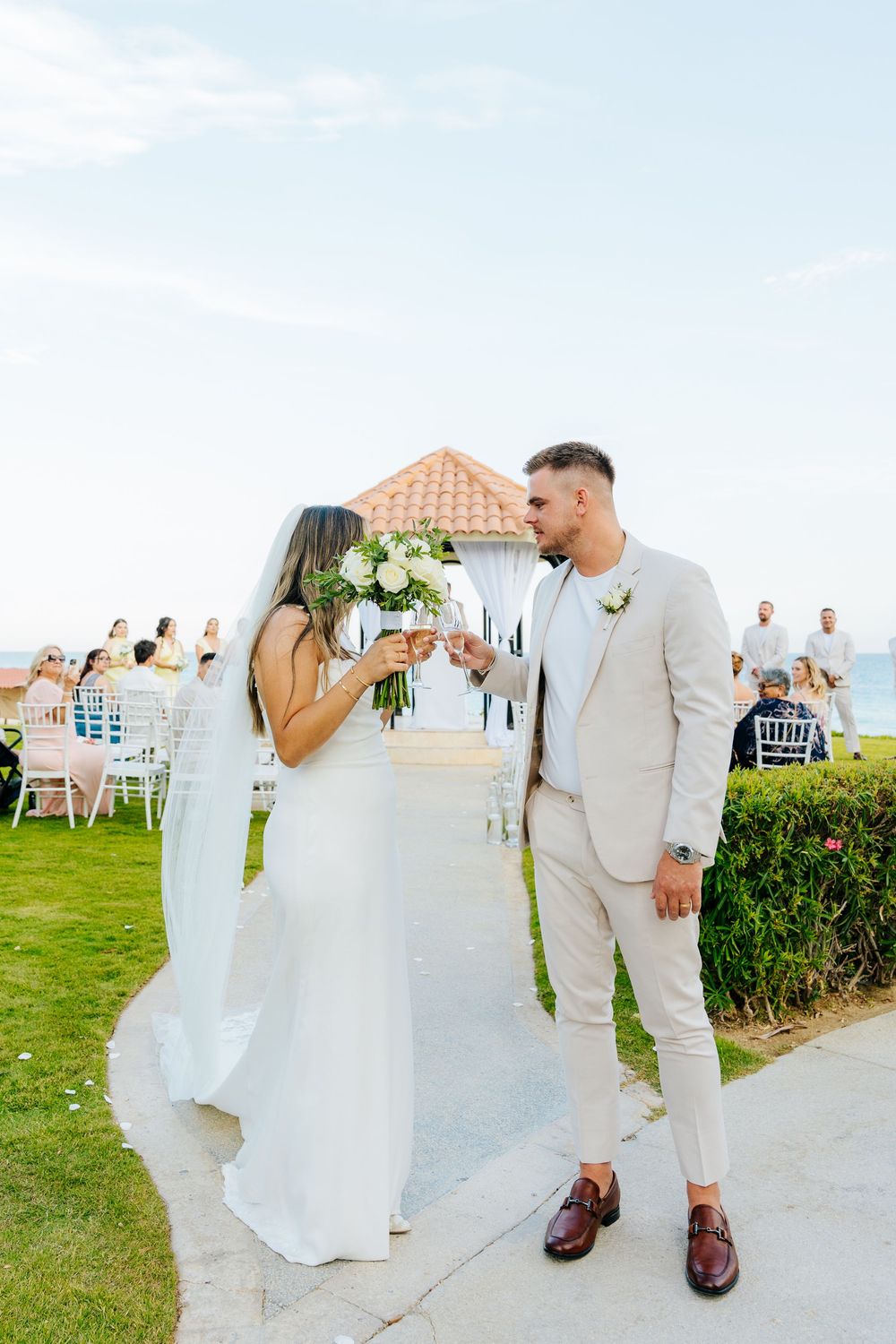 Bride and groom toast with champagne glasses at an outdoor seaside wedding ceremony, with guests seated near a gazebo.