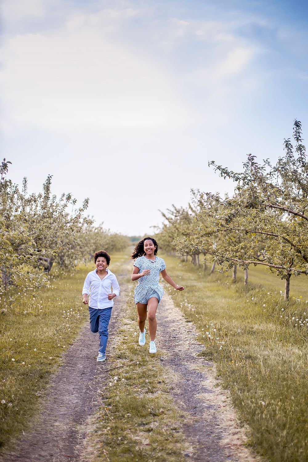 Two people running joyfully down a dirt path between blooming fruit trees in an orchard on a sunny spring day.