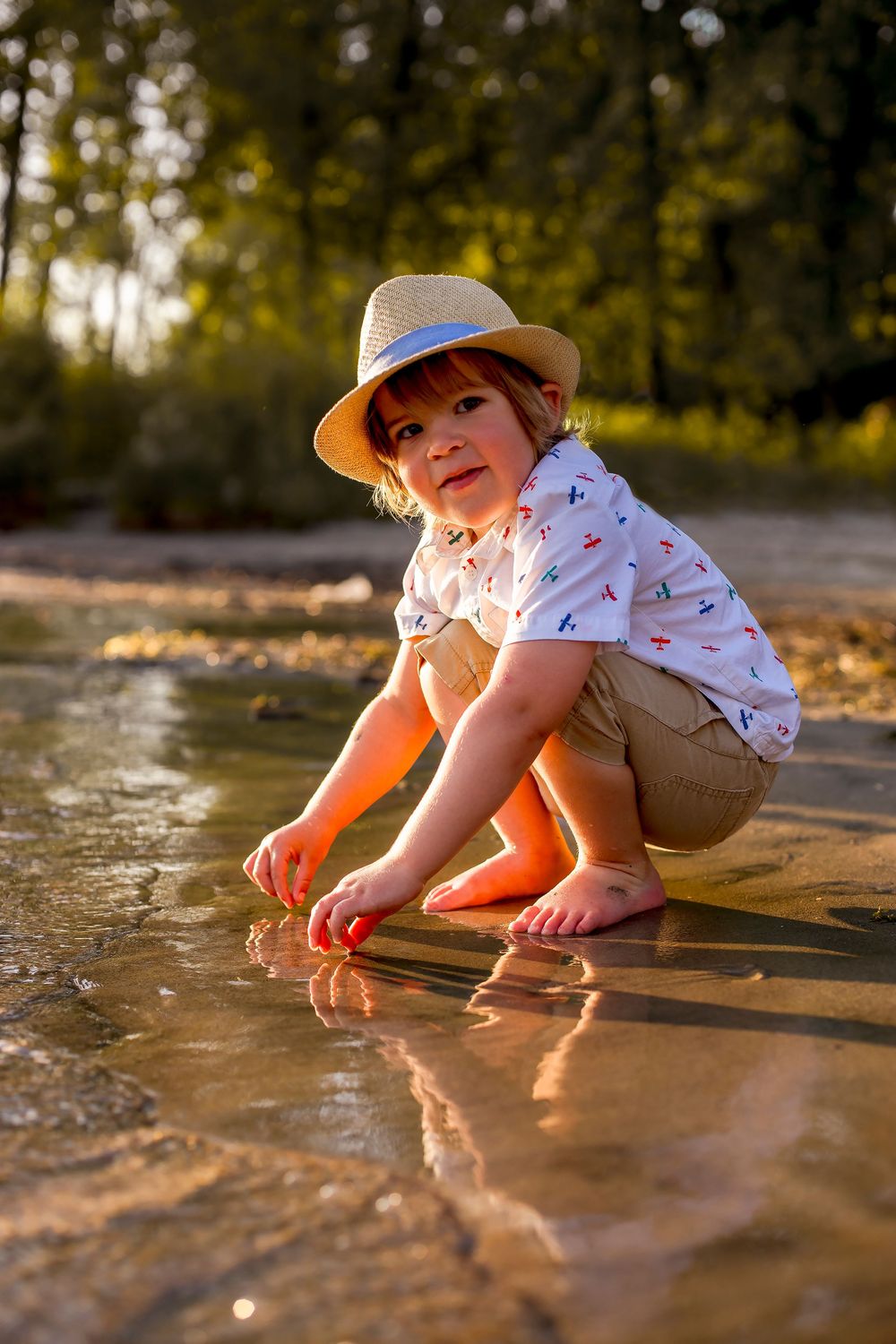 A young child in a straw hat crouches by a shallow river, playing in the water during golden hour.