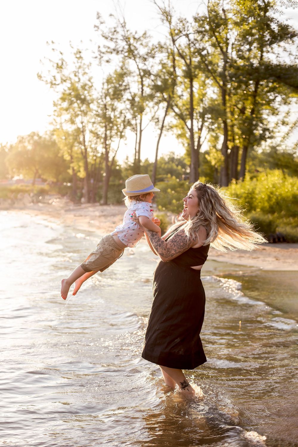 A tattooed mother lifts her baby boy on a sunny beach at golden hour, both smiling near gentle waves and lush trees.