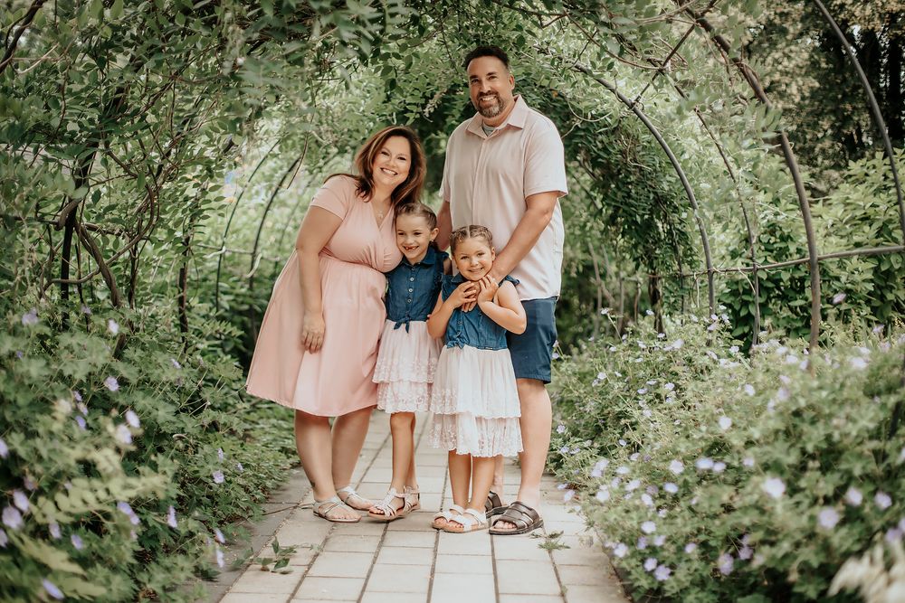 Family of four posing under a lush green garden archway, parents with two young daughters in coordinating outfits.