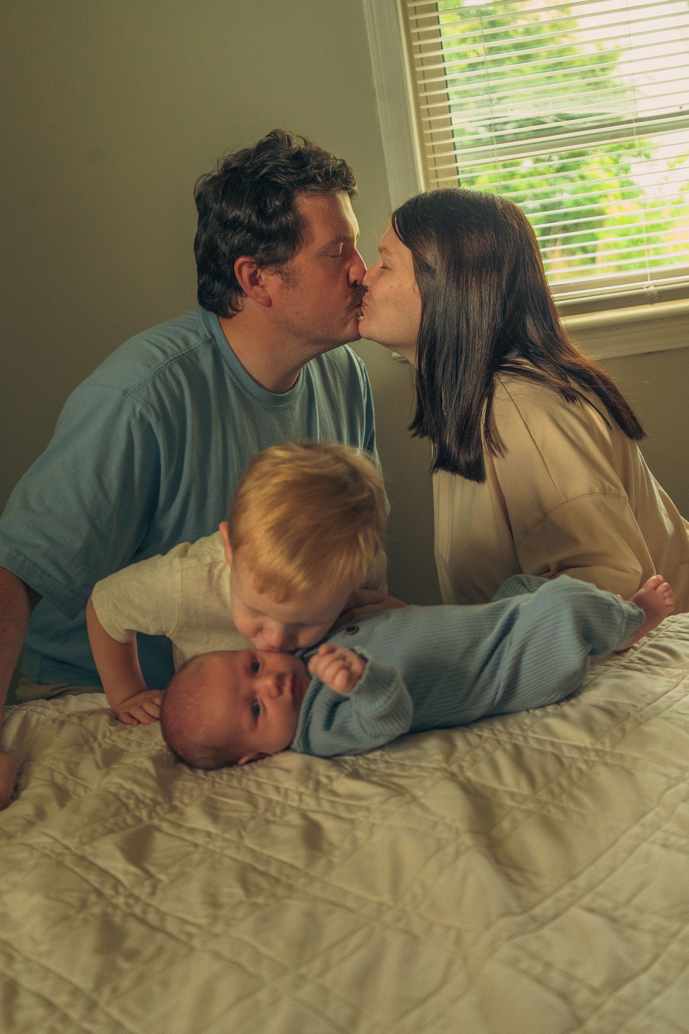 Parents kissing on a bed while their toddler and newborn baby rest between them.