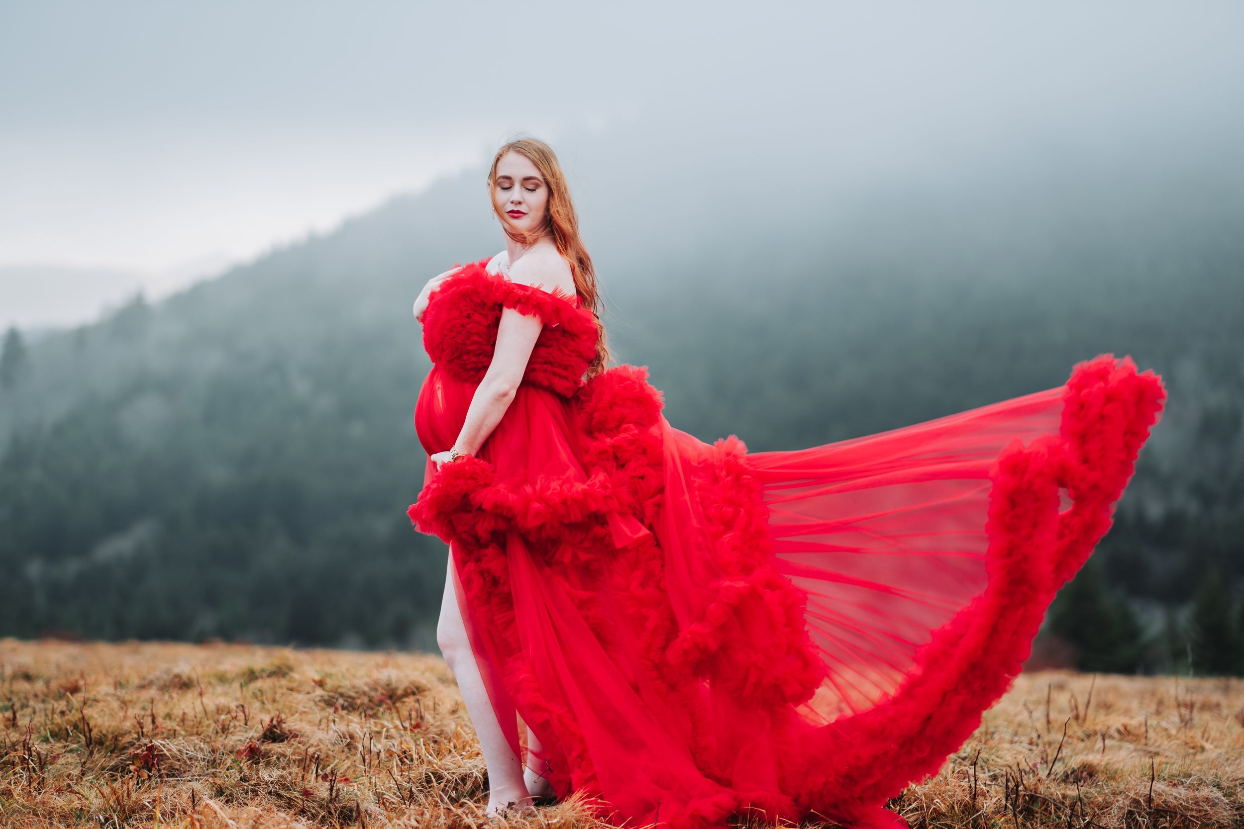 Woman in flowing red tulle gown poses dramatically on misty mountain hillside with dress billowing in wind.