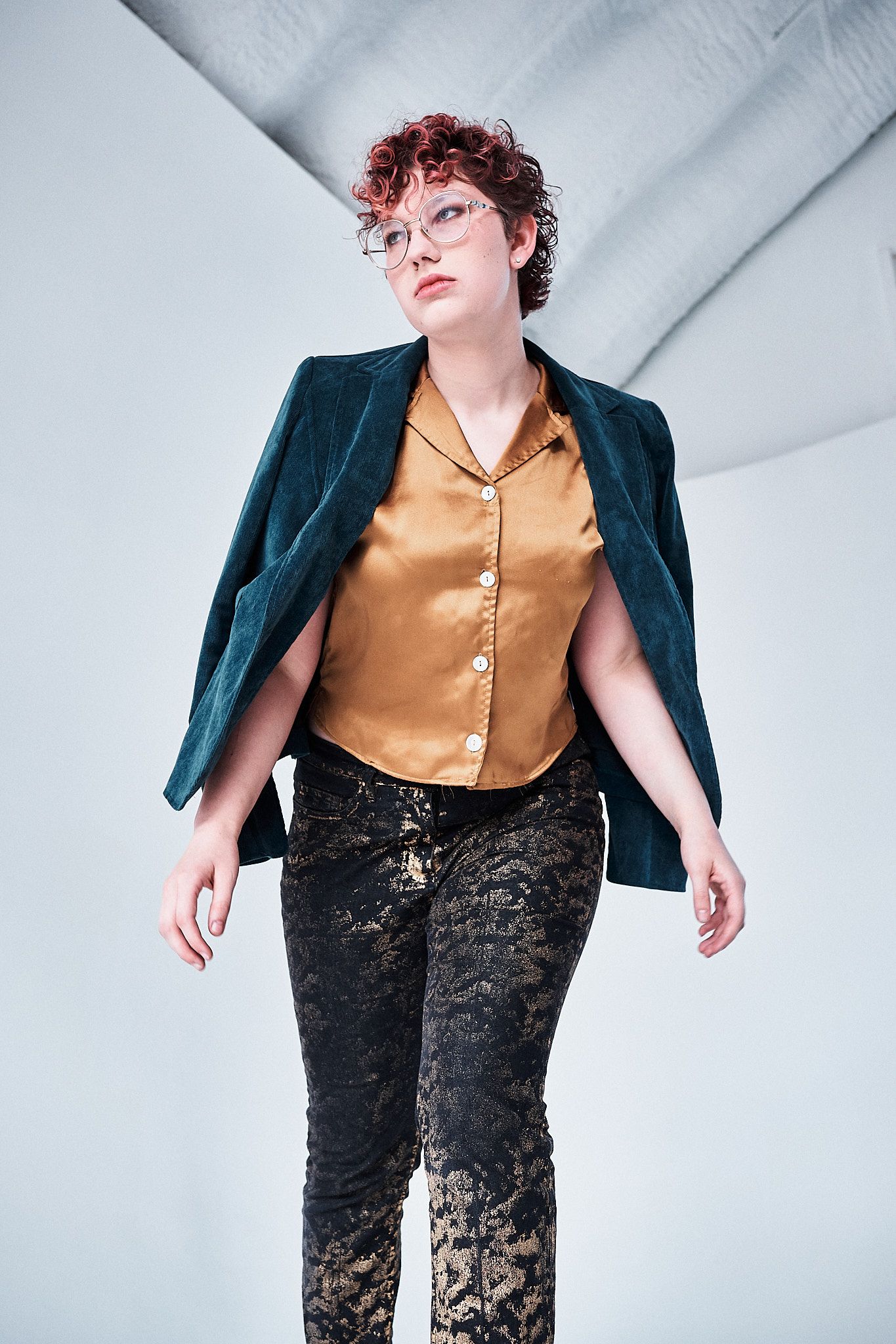 A model with curly red hair and glasses wears a gold satin blouse, dark velvet blazer, and black brocade trousers.