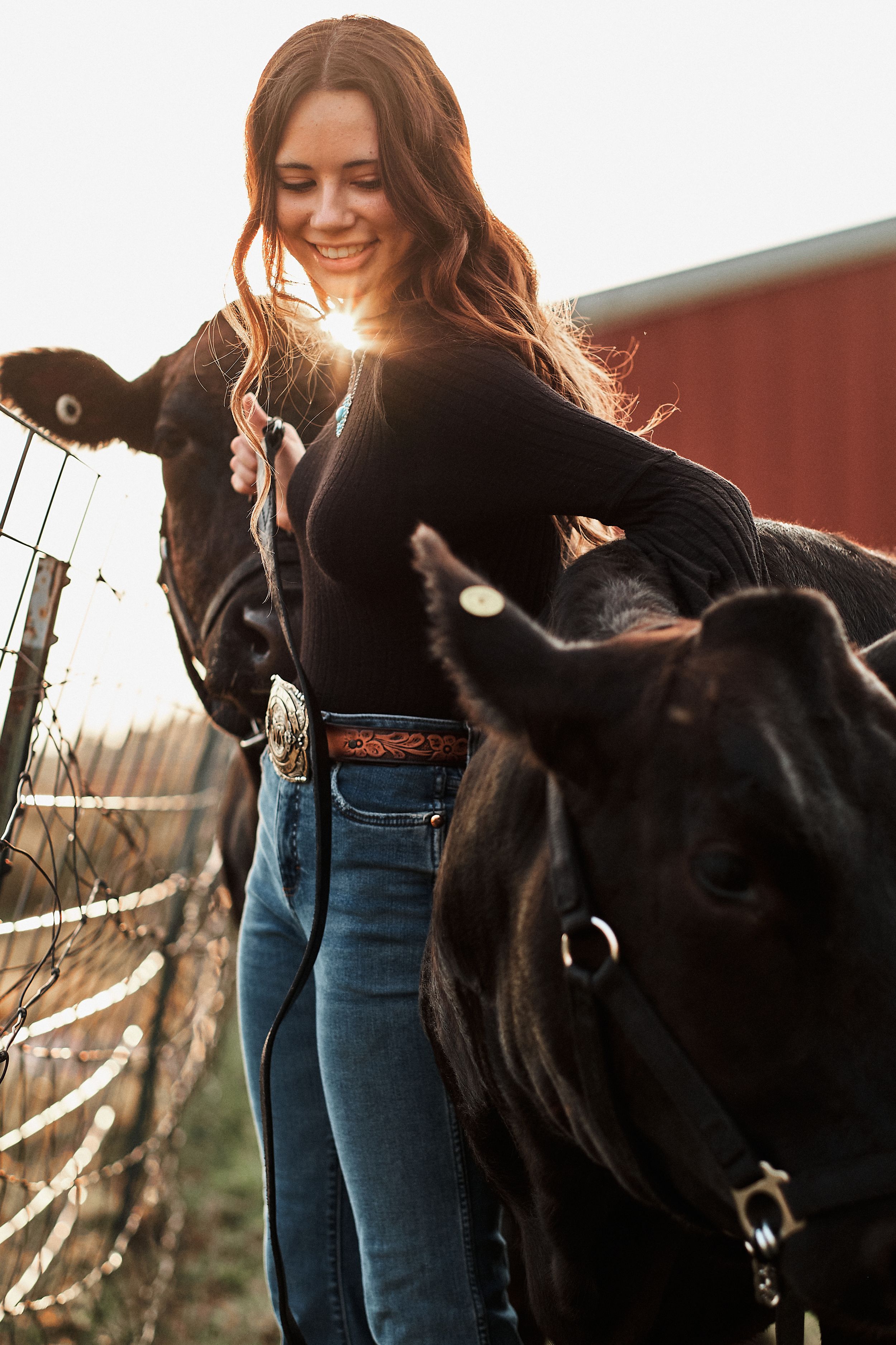 Young woman in black top and jeans smiles while standing with black cattle near a red barn at sunset.
