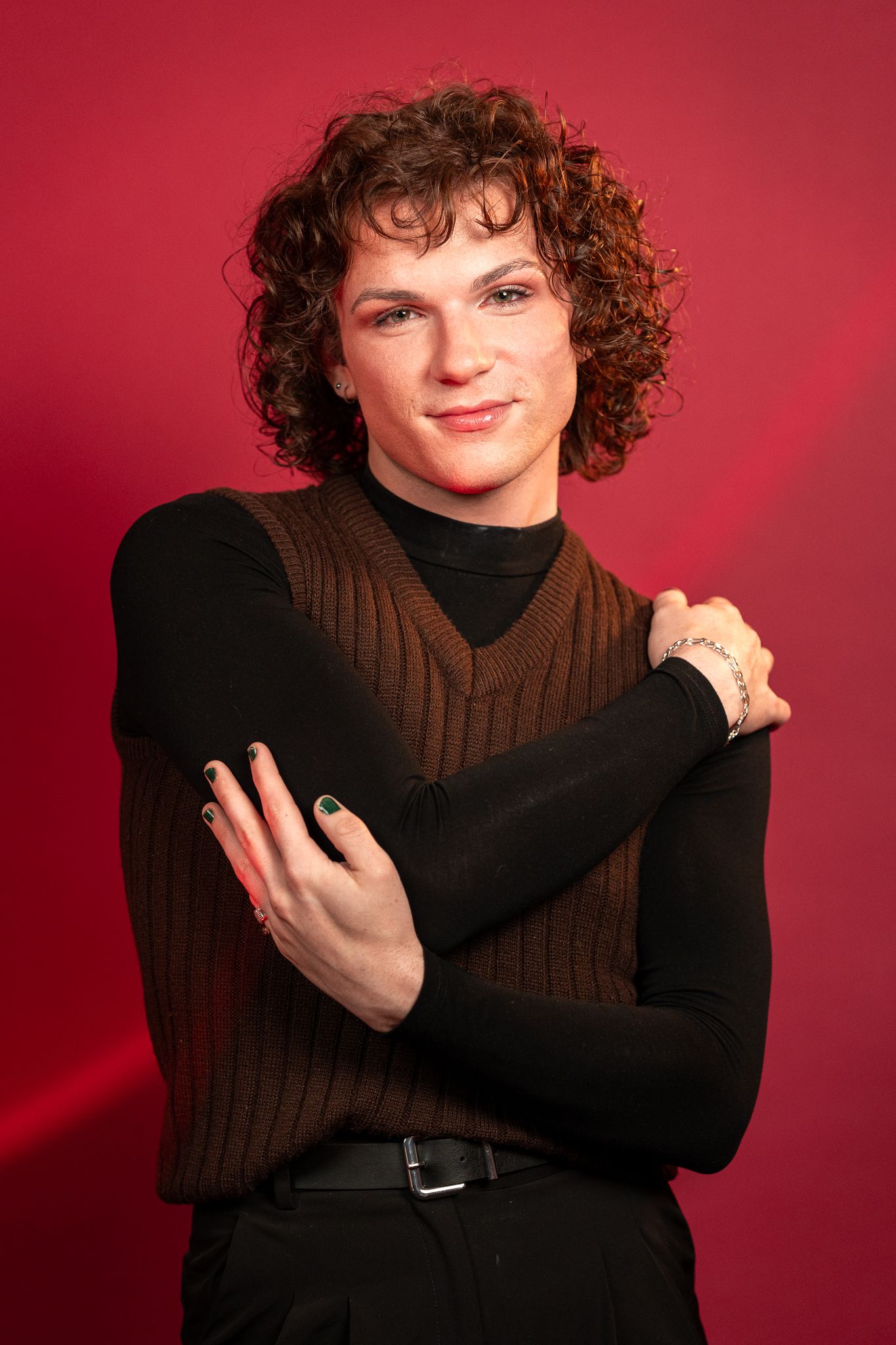 Person with curly hair in brown vest and black turtleneck poses with crossed arms against red background.