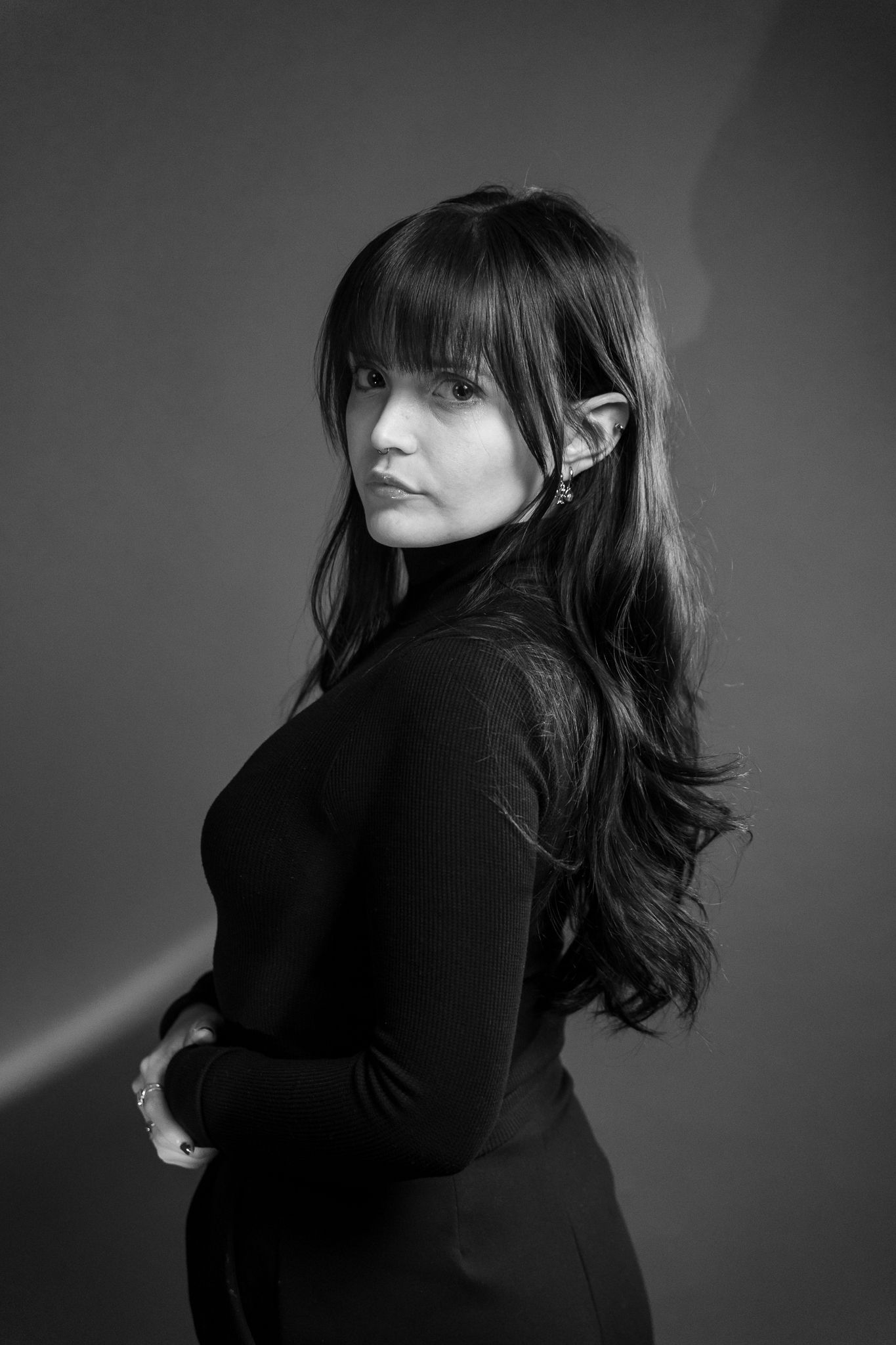 Black and white portrait of a young woman with long dark hair and bangs, wearing a black turtleneck, looking over shoulder.