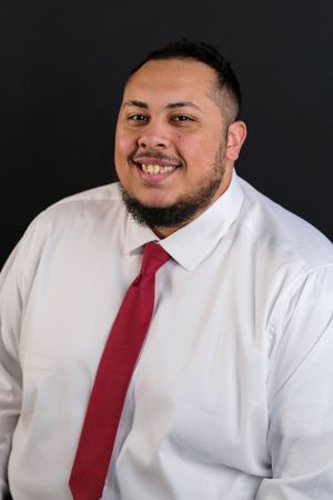 Smiling man with beard wearing white dress shirt and red tie posed against dark background for professional headshot.