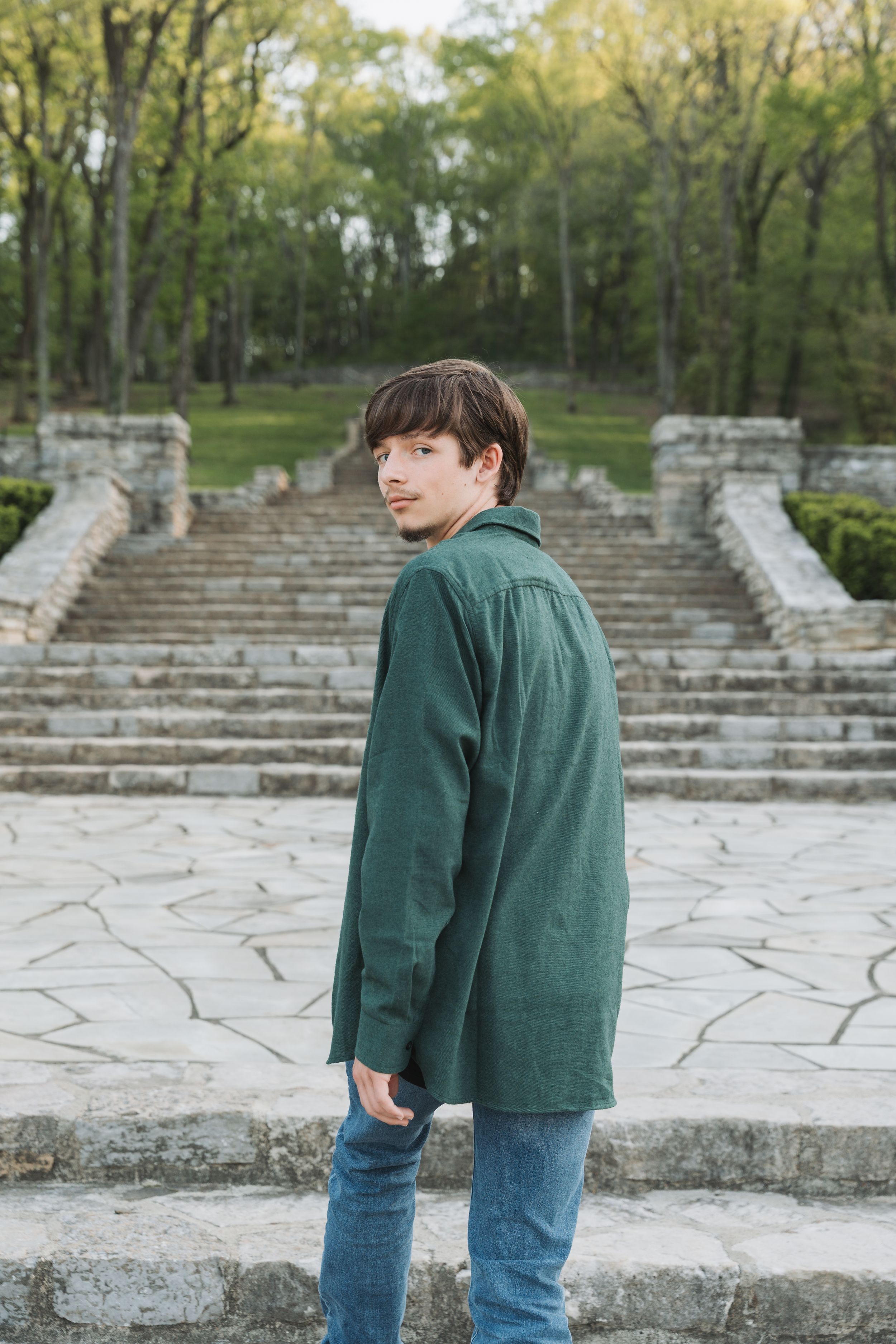 Young man in green shirt and jeans standing on stone steps in a park, looking back over his shoulder.