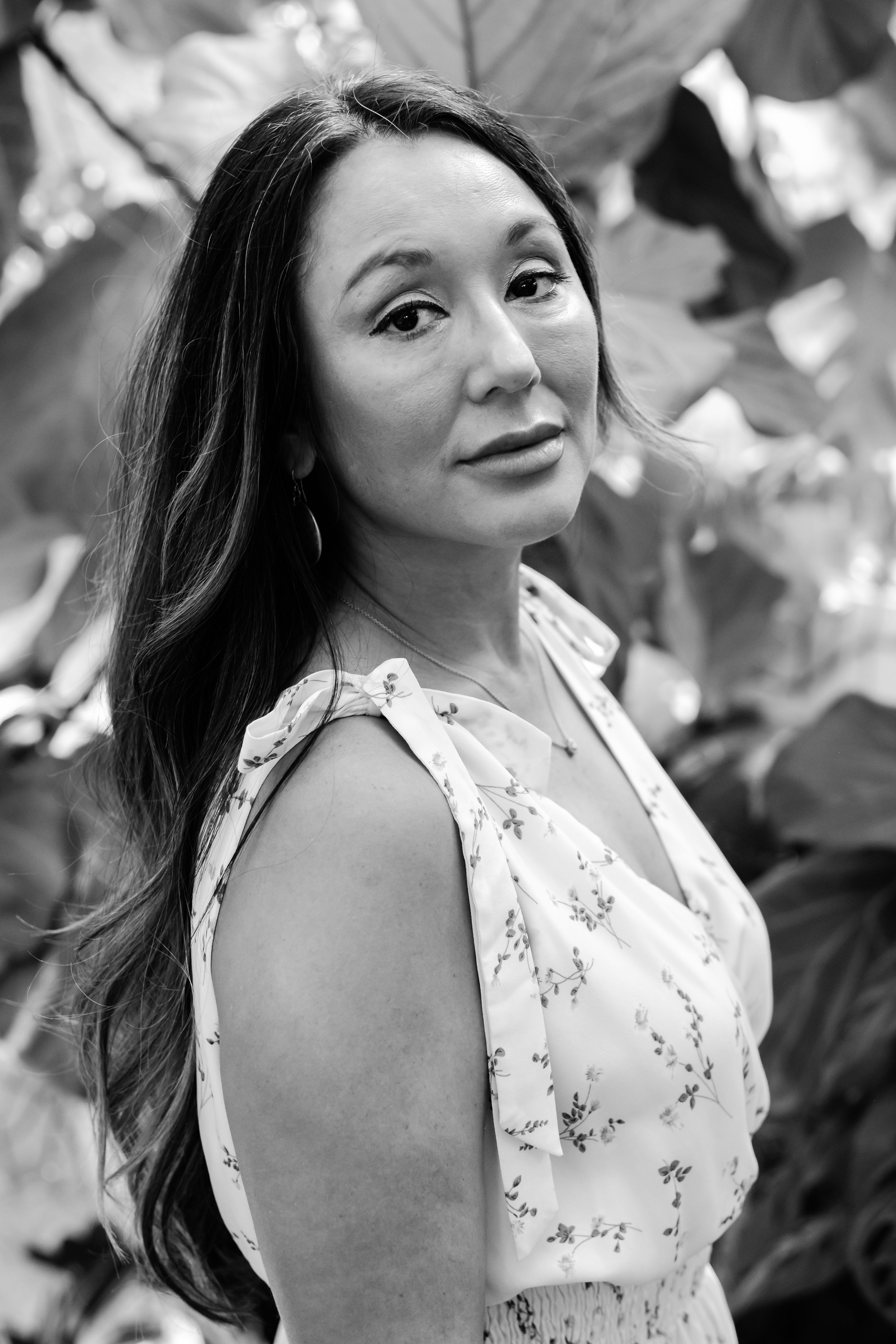 Black and white portrait of a woman in a floral dress standing among large leaves outdoors.
