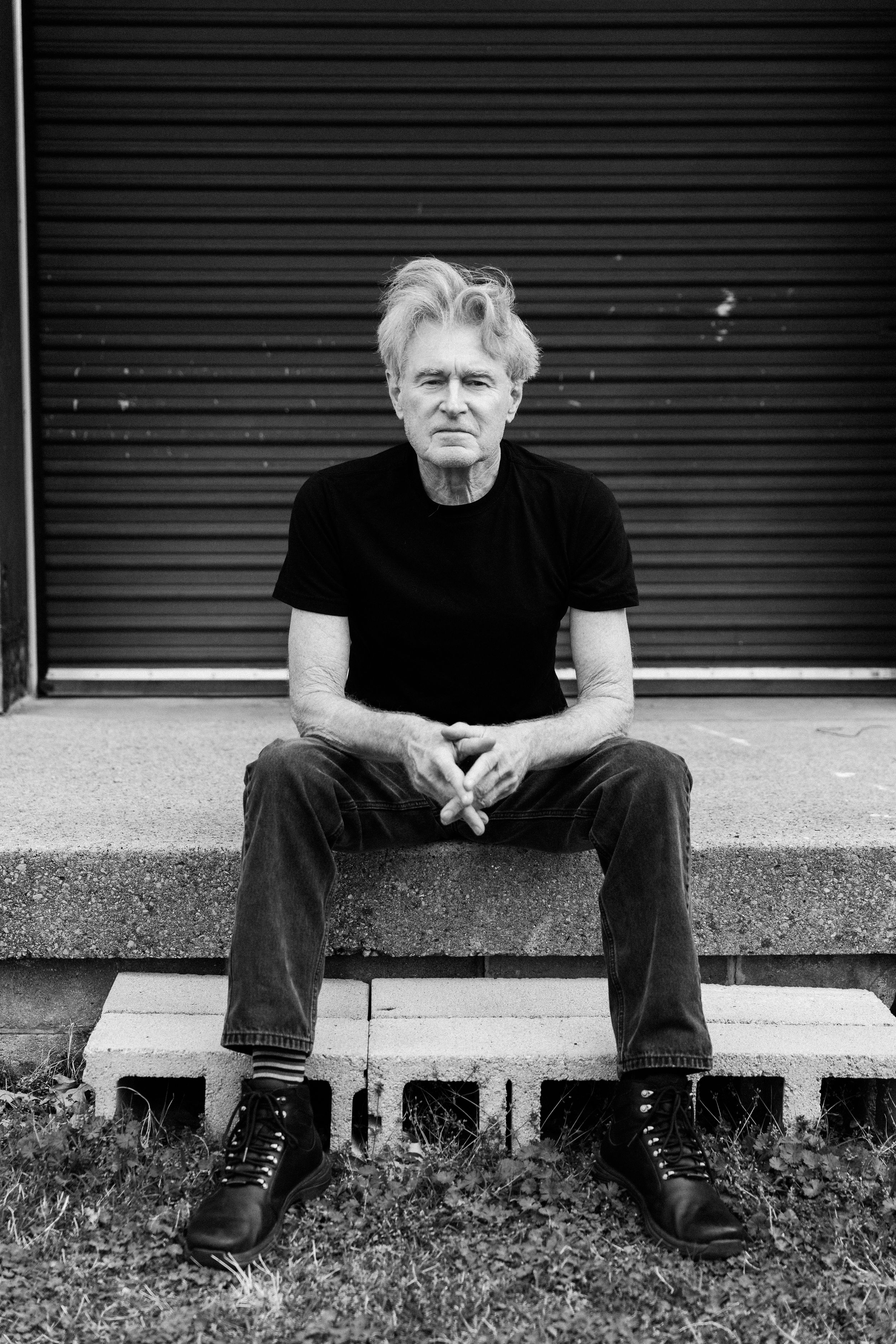 Black and white portrait of an older man with gray hair sitting on steps in a black t-shirt and jeans.