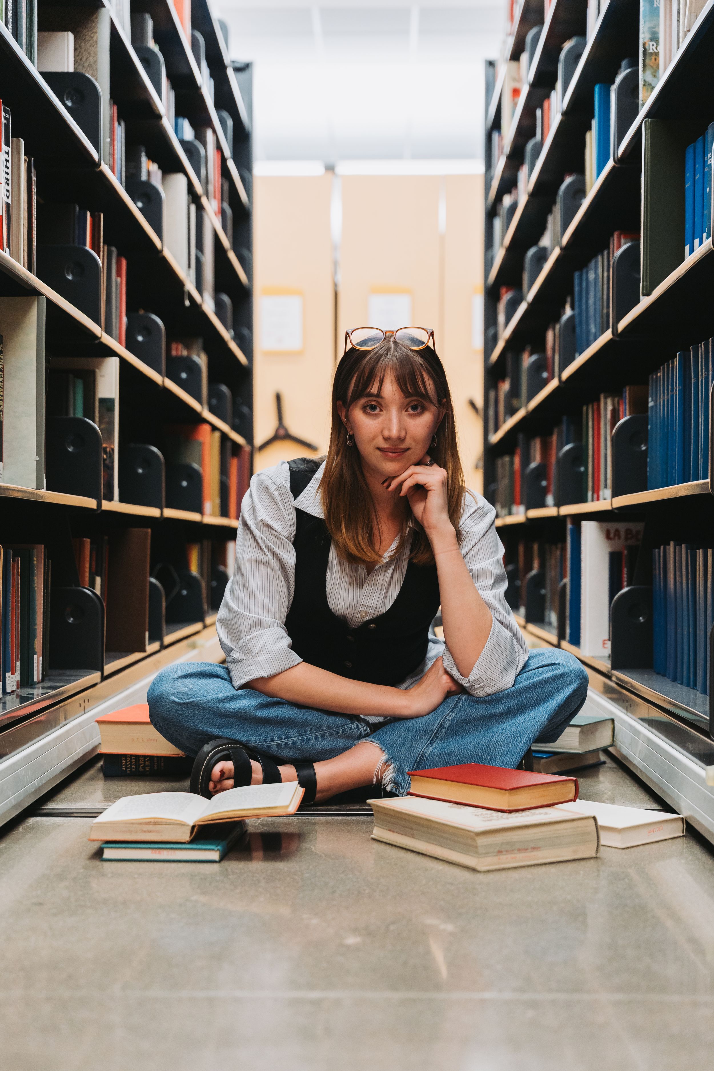 Young woman sitting cross-legged on library floor surrounded by books, wearing glasses on head, smiling at camera.