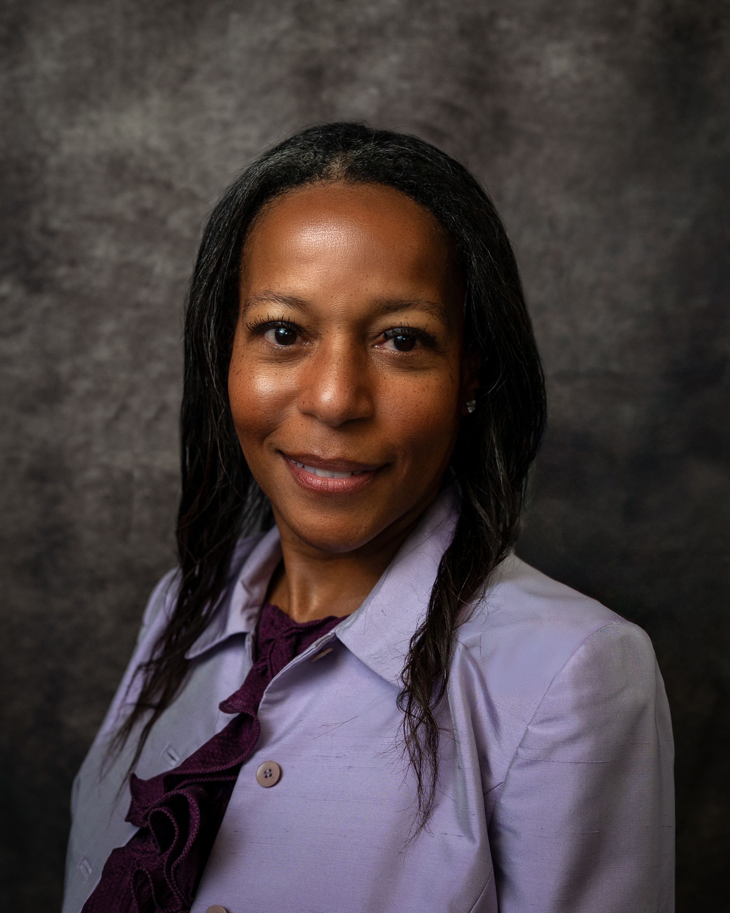 Professional headshot of a woman with long dark hair wearing a lavender blazer against a gray backdrop.