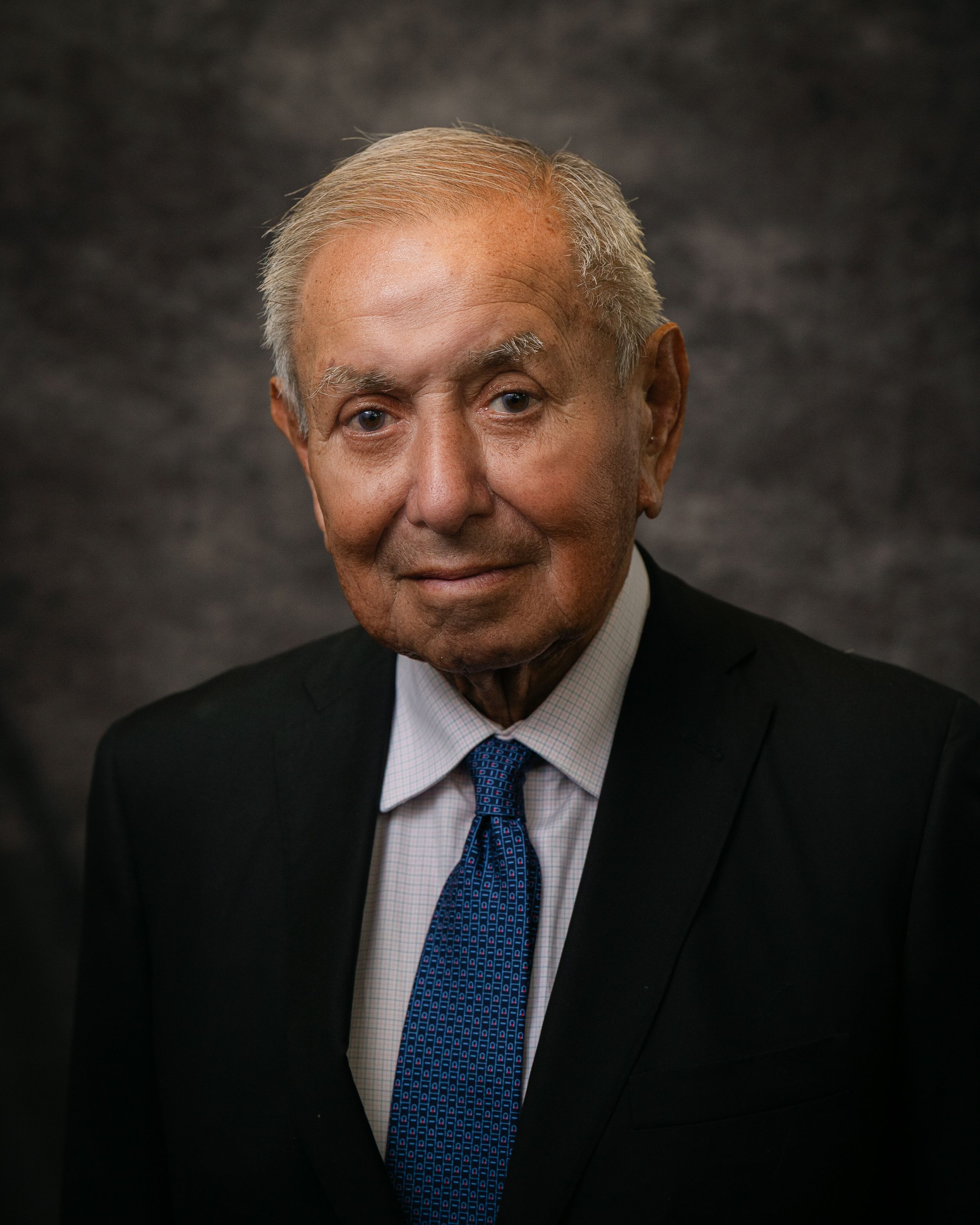Professional headshot of an elderly man in a black suit and blue tie, smiling against a gray studio background.