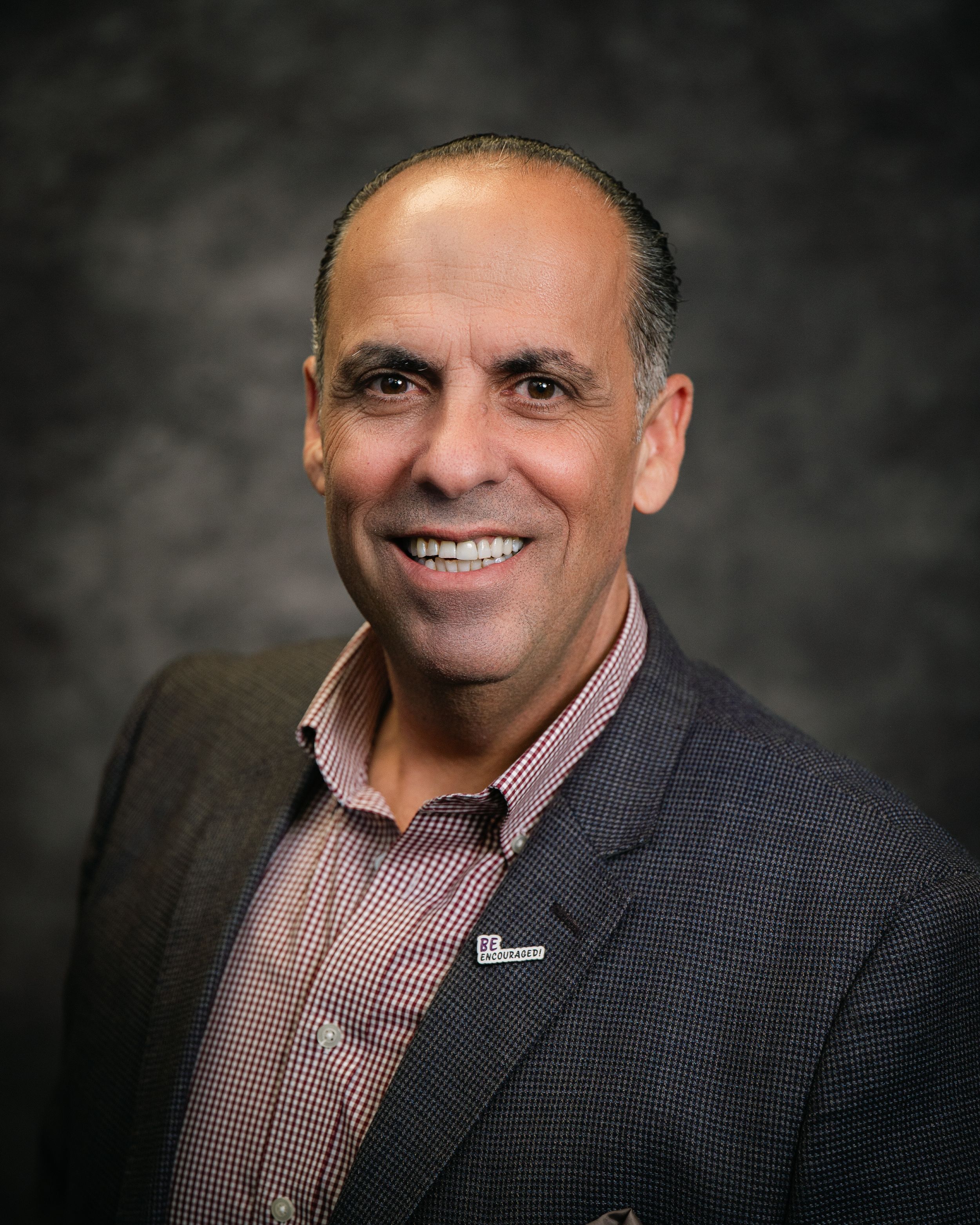 Professional headshot of a smiling middle-aged man in a dark blazer and checkered shirt against a gray background.