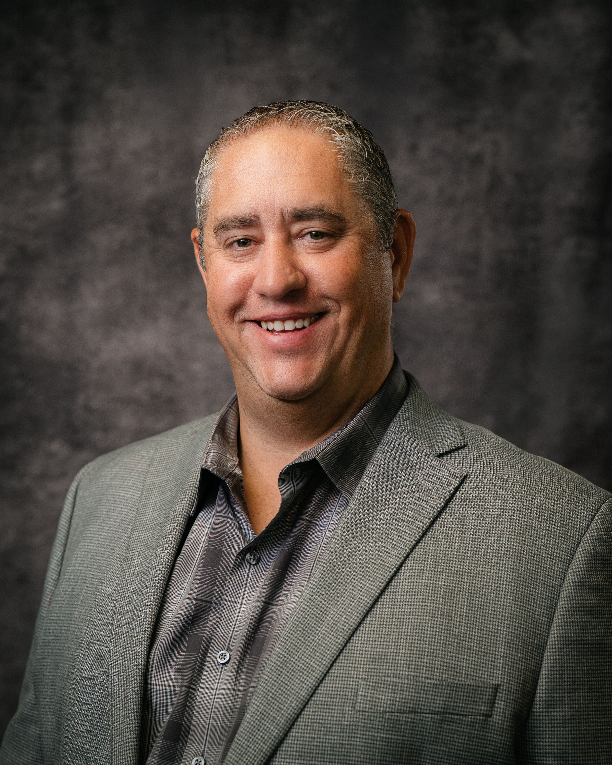 Professional headshot of a smiling middle-aged man in a gray blazer and plaid shirt against a dark studio background.