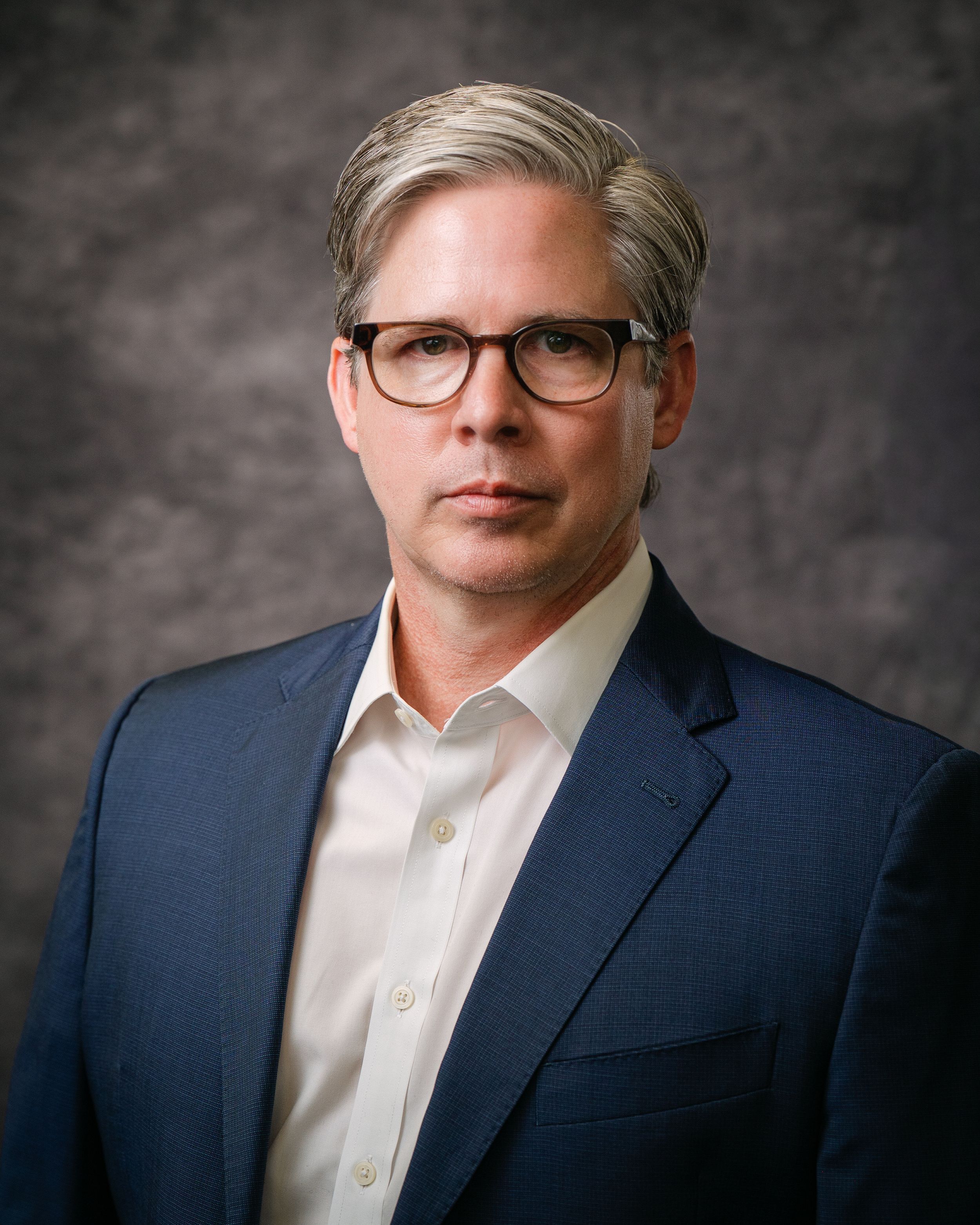 Professional headshot of a middle-aged man in a blue blazer and glasses with gray-streaked hair on a neutral background.