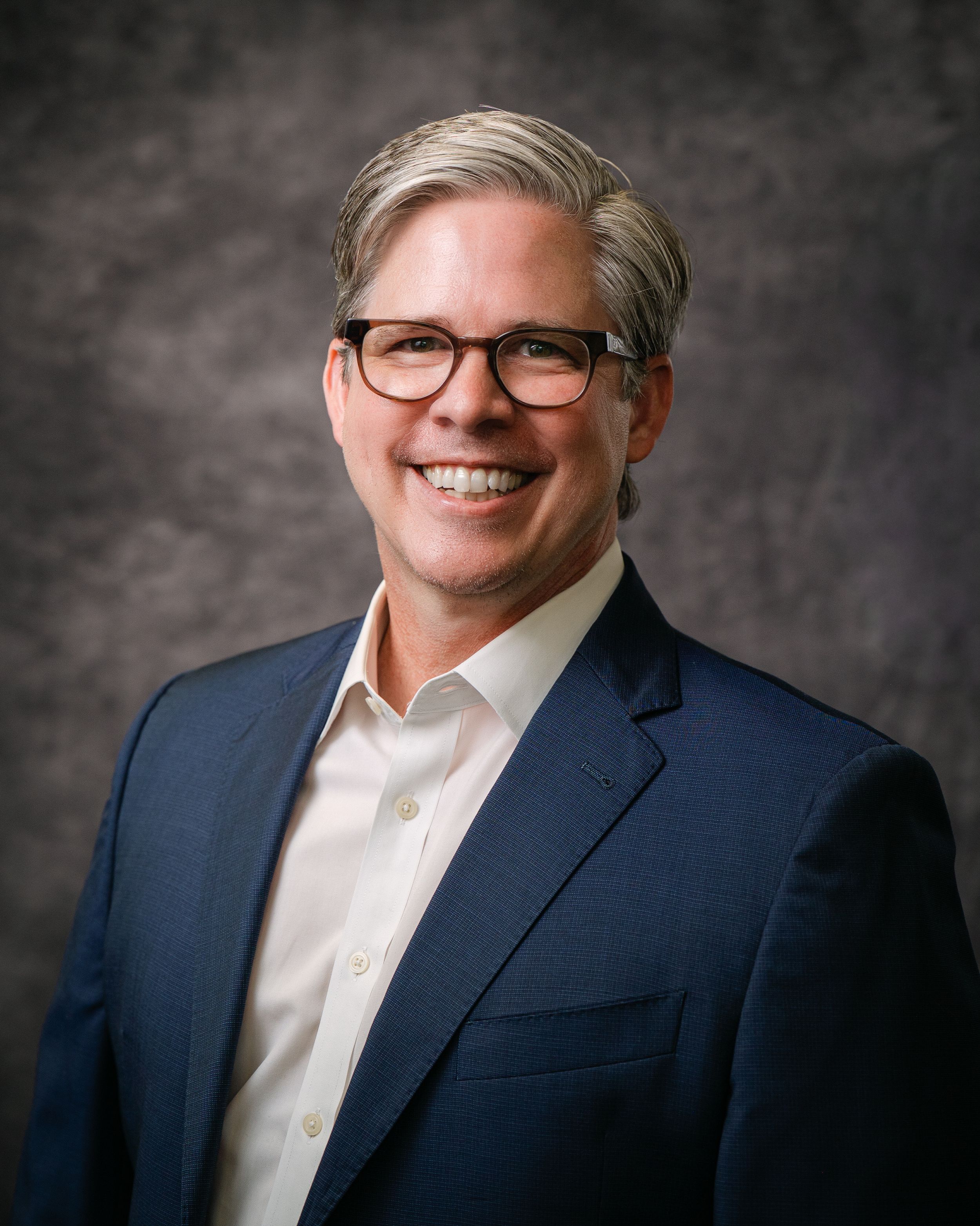 Professional headshot of a smiling middle-aged man with gray hair and glasses wearing a navy blue blazer.