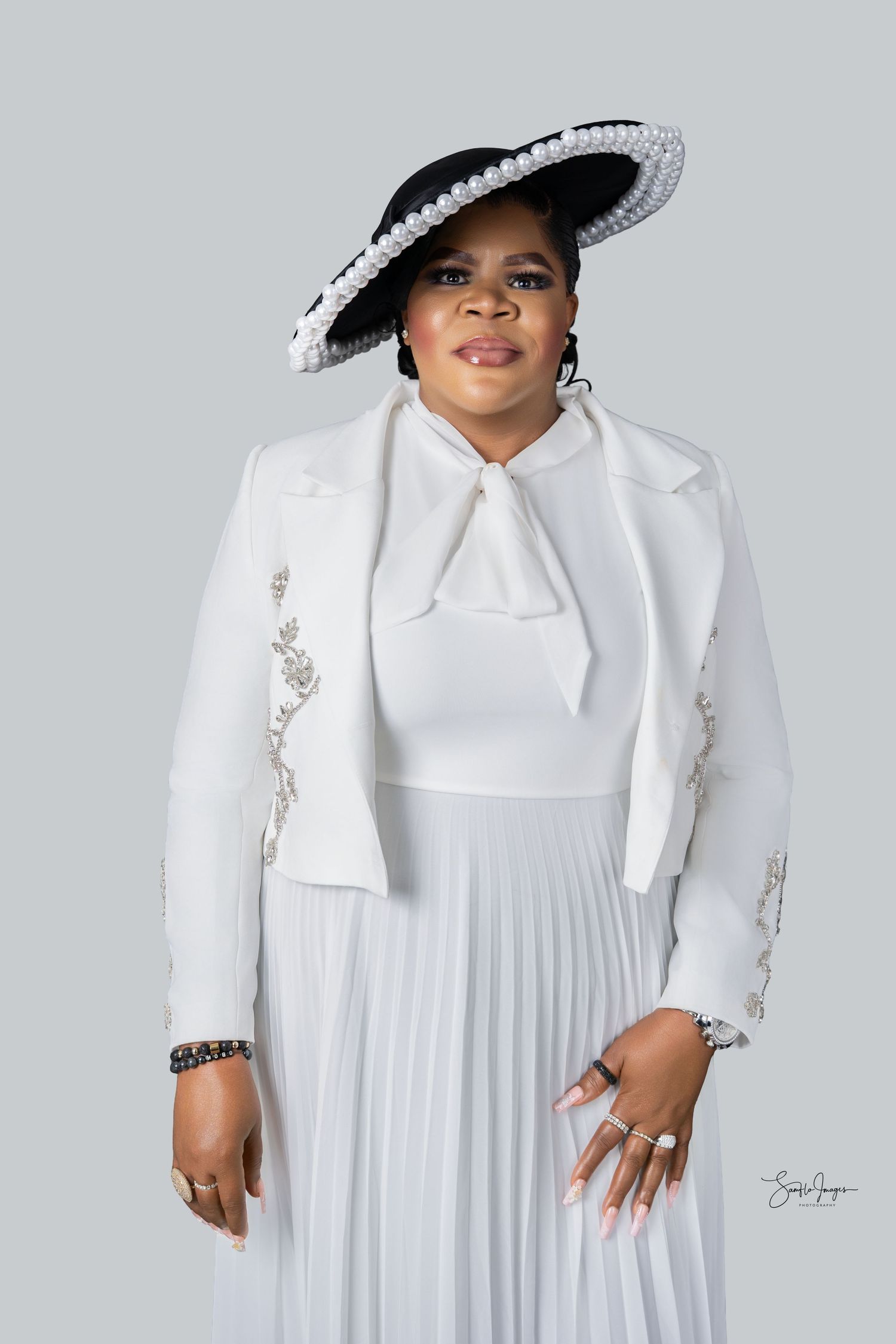 Woman in elegant white pleated dress, lace jacket, and black pearl-trimmed hat poses for a formal portrait.