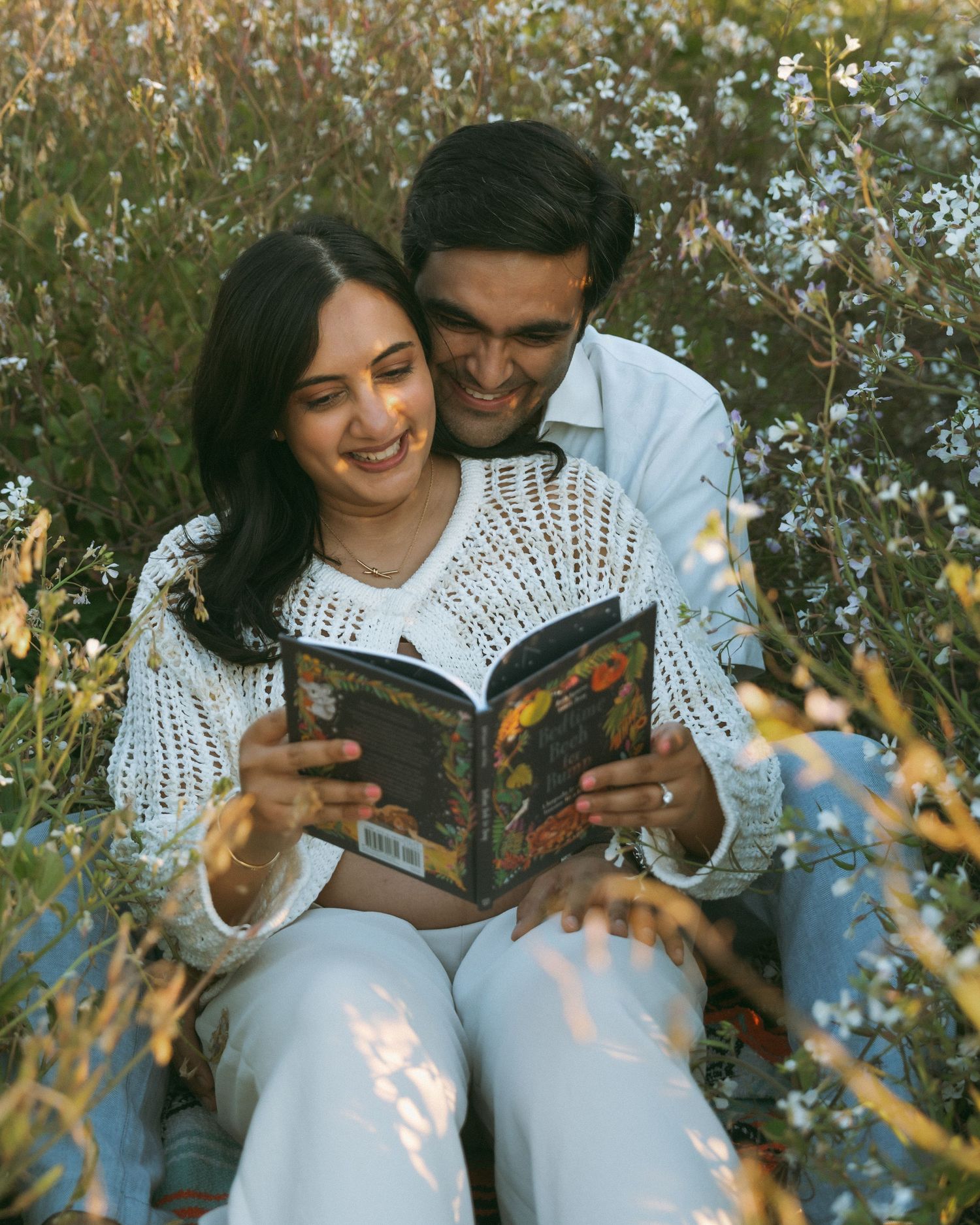 A smiling couple sits together in a wildflower field, sharing a book in warm golden sunlight.