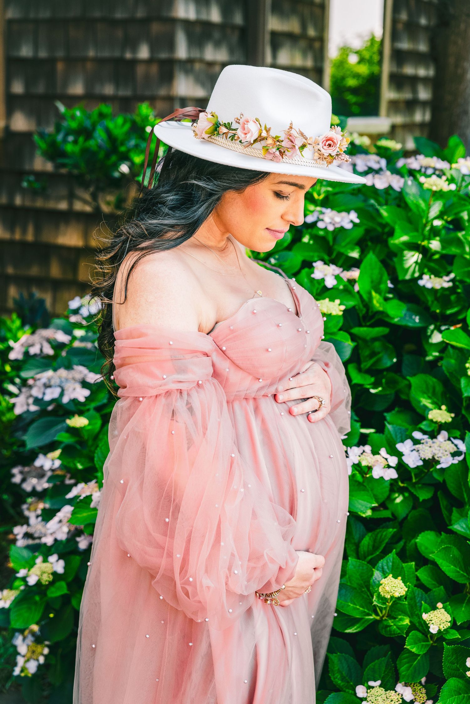 Pregnant woman in pink tulle off-shoulder gown and floral white hat poses near blooming hydrangeas.