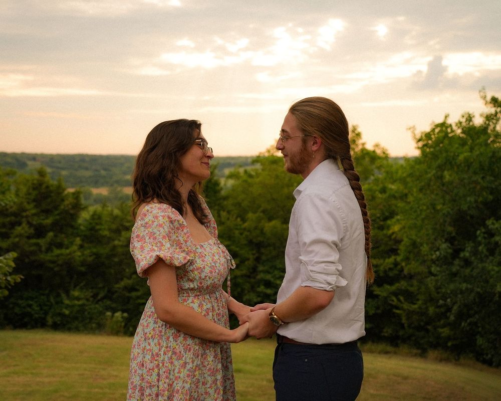 Romantic couple holding hands facing each other on a grassy hill at golden hour with lush trees behind.