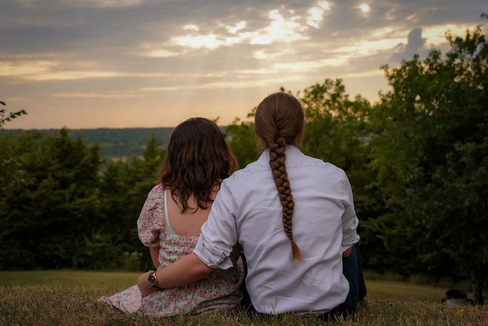 A couple sits together on a grassy hill watching a beautiful sunset over a lush green landscape.
