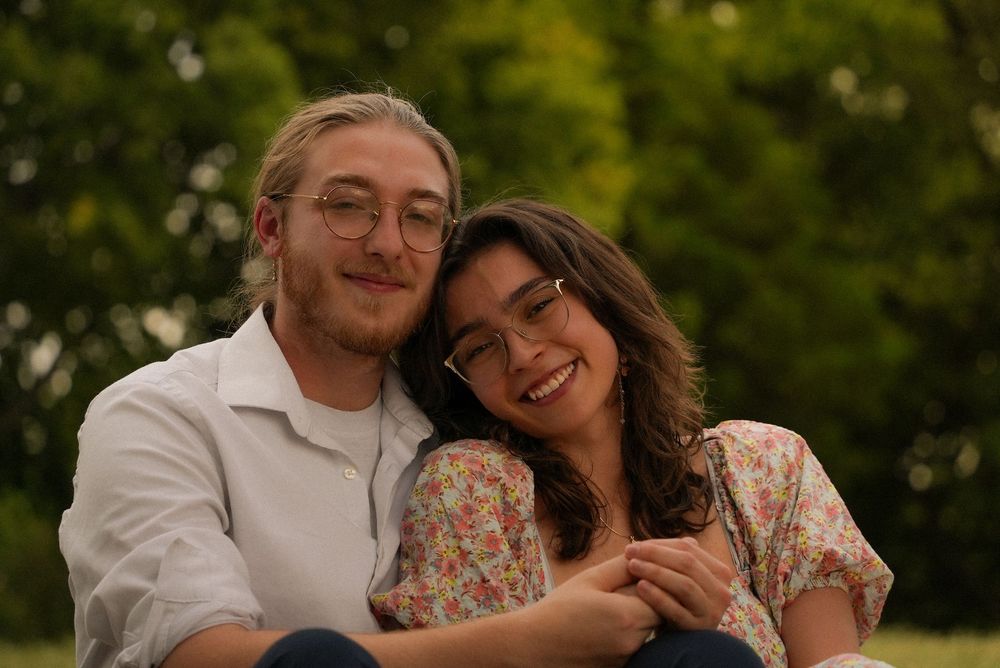 A happy young couple with glasses sit together smiling in a lush green park setting.