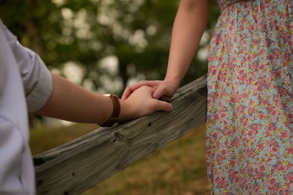 Couple holding hands over a wooden fence rail in a lush outdoor setting.