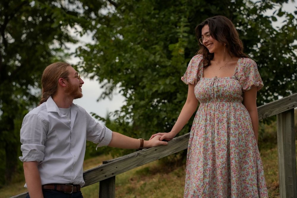 Romantic couple holding hands on a wooden fence surrounded by lush green trees outdoors.