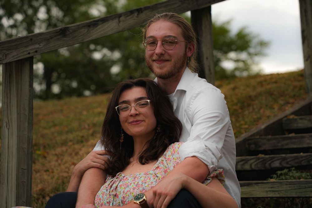 A young couple sitting on wooden steps outdoors, smiling warmly, woman wearing glasses and floral top.