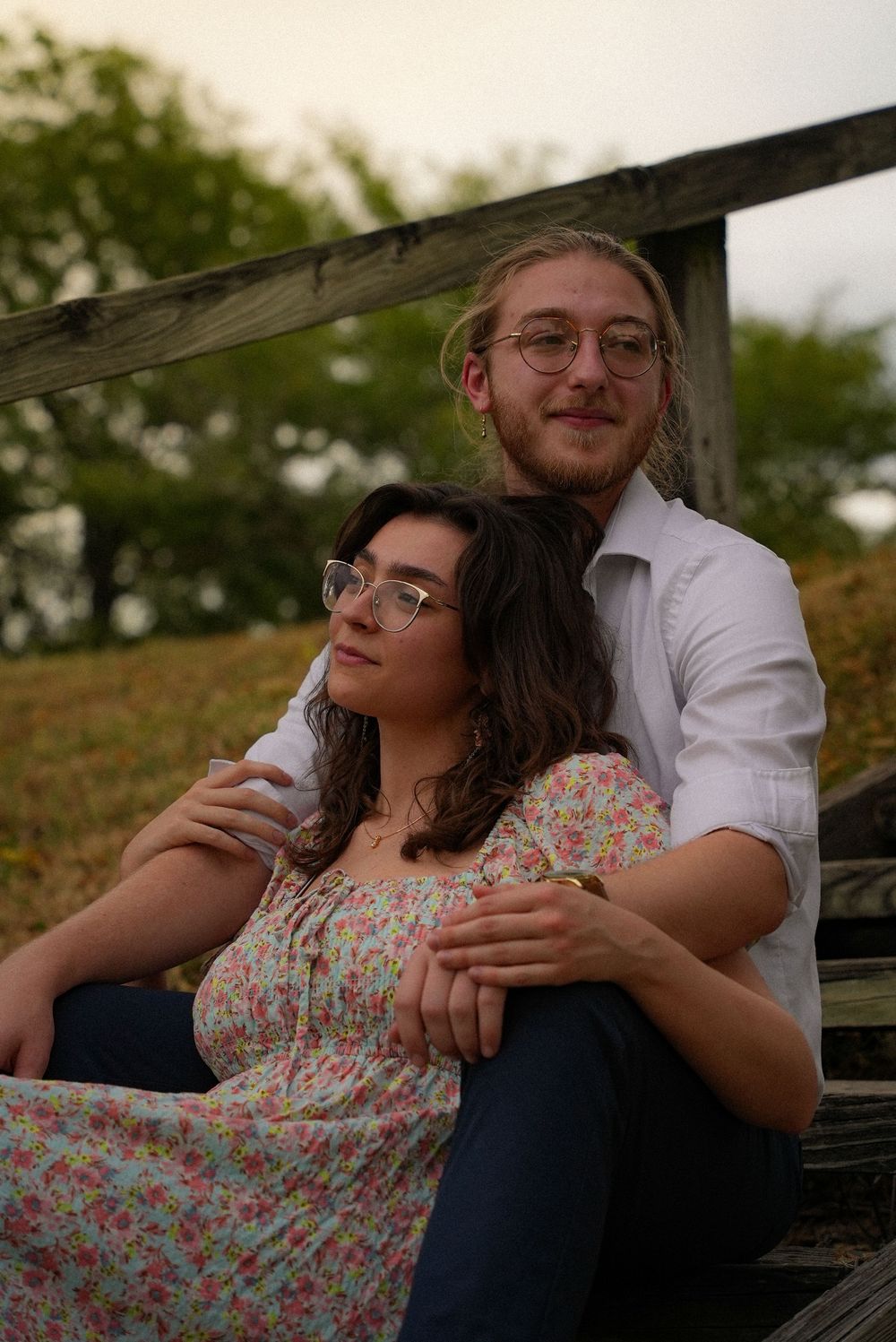 A smiling couple sits on wooden steps outdoors, the woman leaning back against the man in a floral dress.