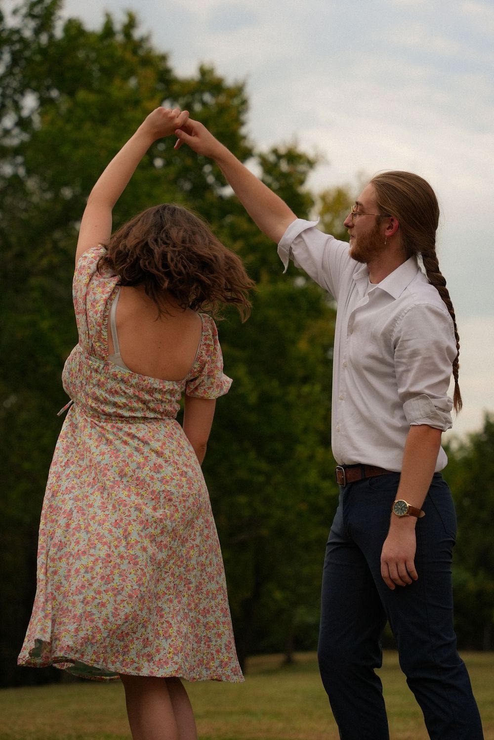 Couple dancing outdoors in a park, woman in floral dress being twirled by long-haired man in white shirt.