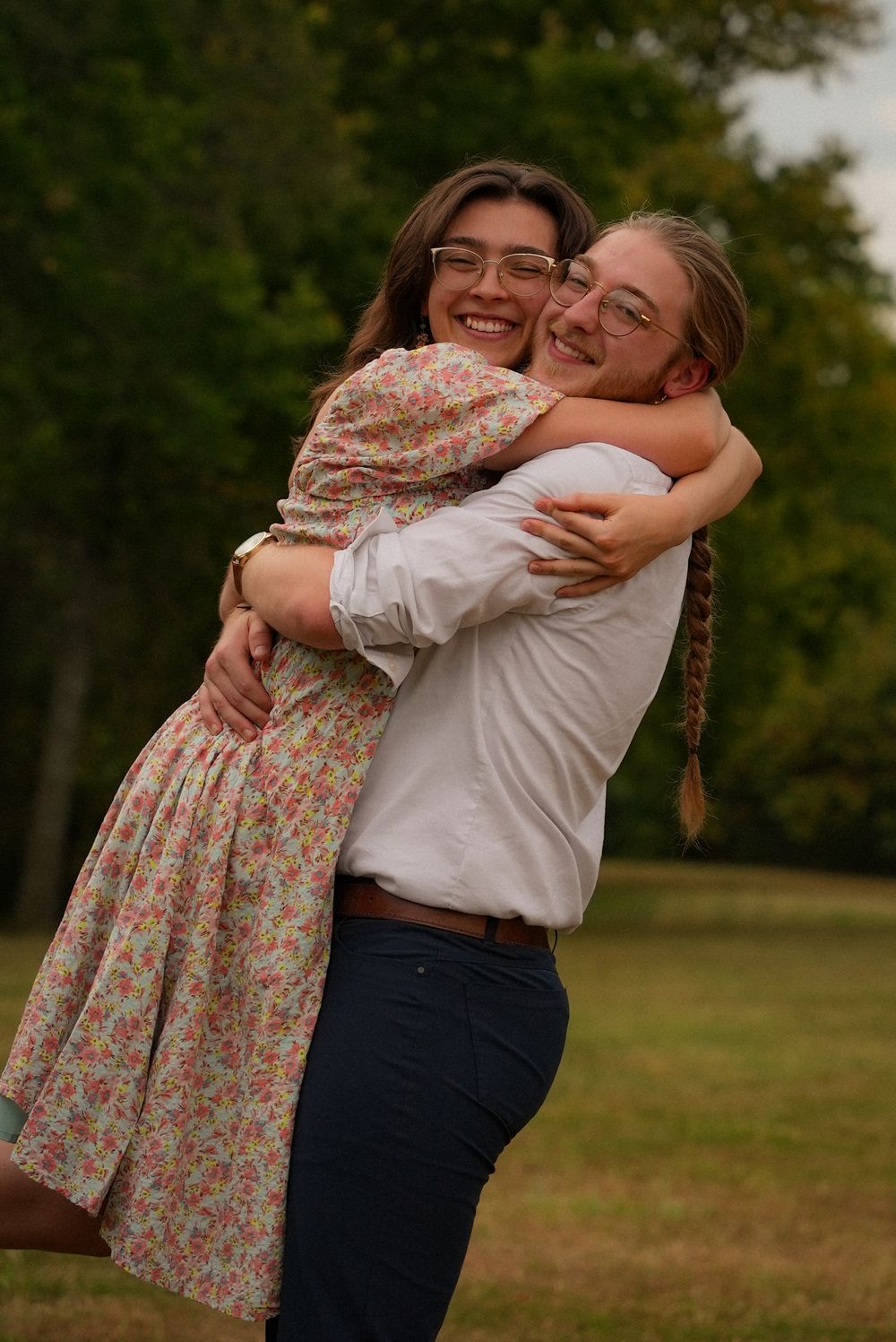 Two smiling young people wearing glasses embrace warmly in a green outdoor park setting.