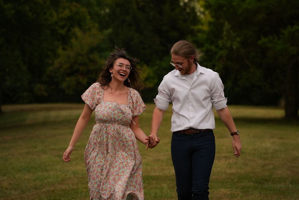 A joyful couple holds hands and laughs while walking together across a lush green park lawn.